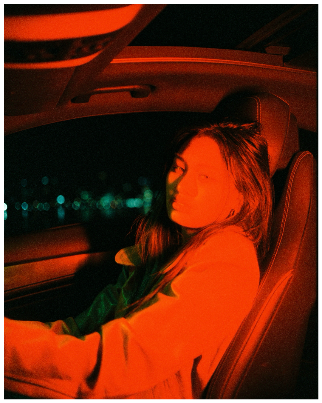 Someone sitting in a car at night illuminated by red lighting with city lights visible in the background.