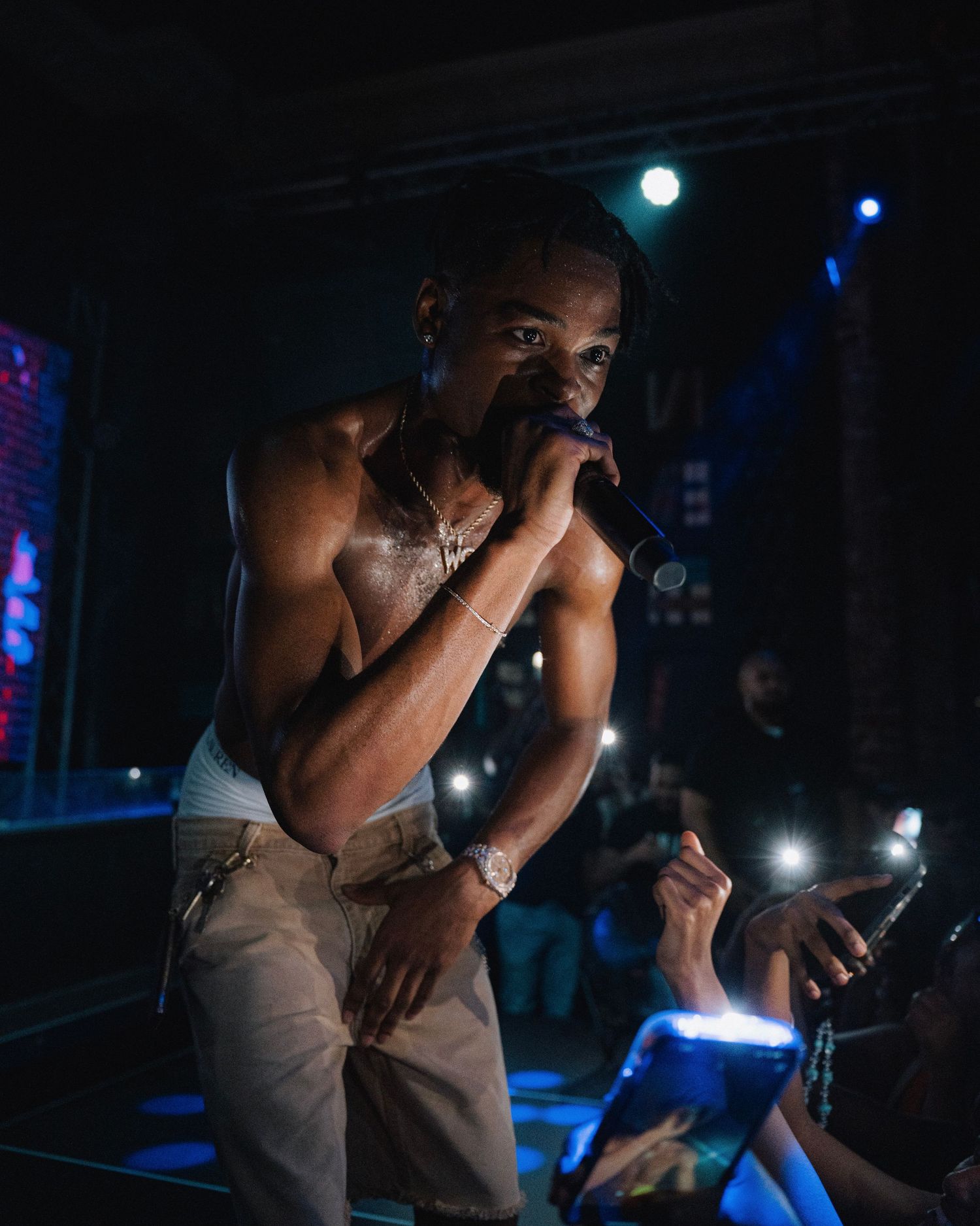 Performer in tank top singing energetically on stage with blue lighting and electronic displays.