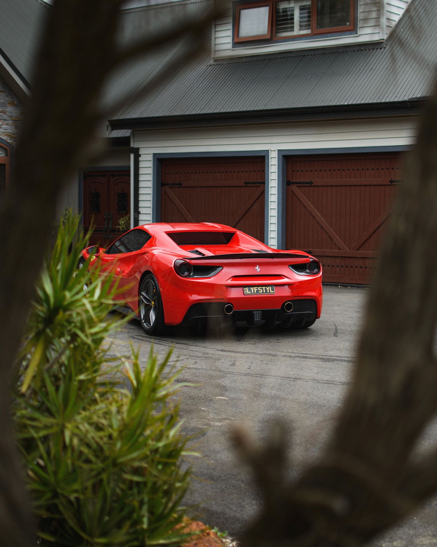 Red Ferrari supercar parked in front of wooden garage doors, viewed through foliage.