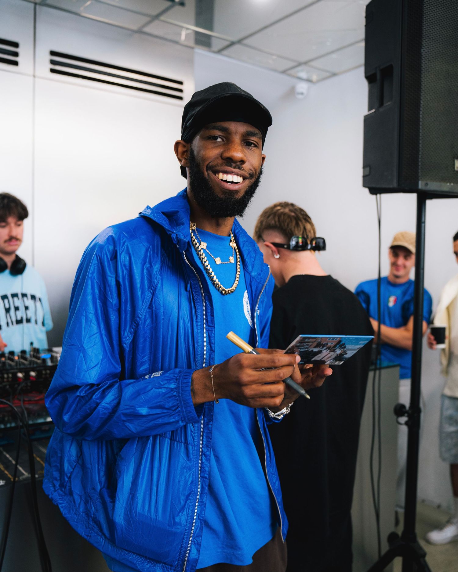 Person wearing bright blue jacket and black cap smiling while holding device at indoor event.