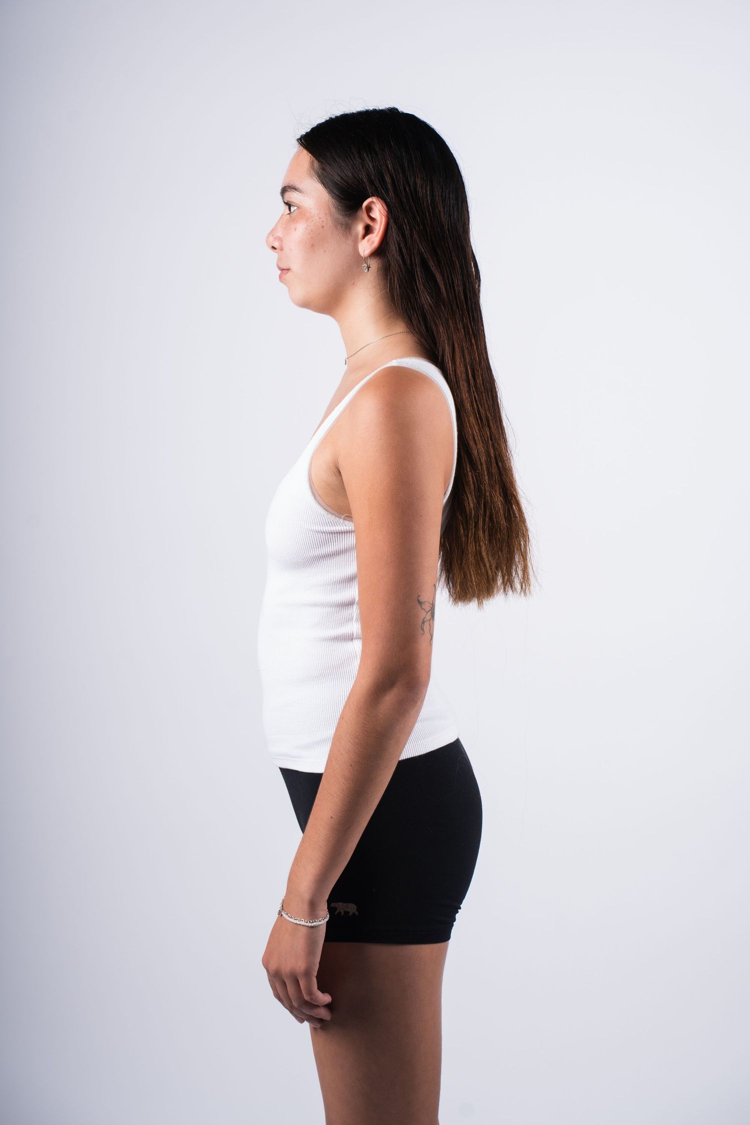 Side profile of person with long dark hair wearing white tank top and black shorts against light background.