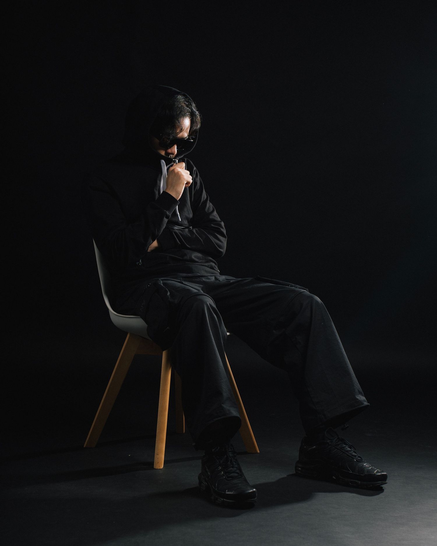 Dramatic dark studio portrait shows figure in black clothing seated on modern chair against black backdrop.