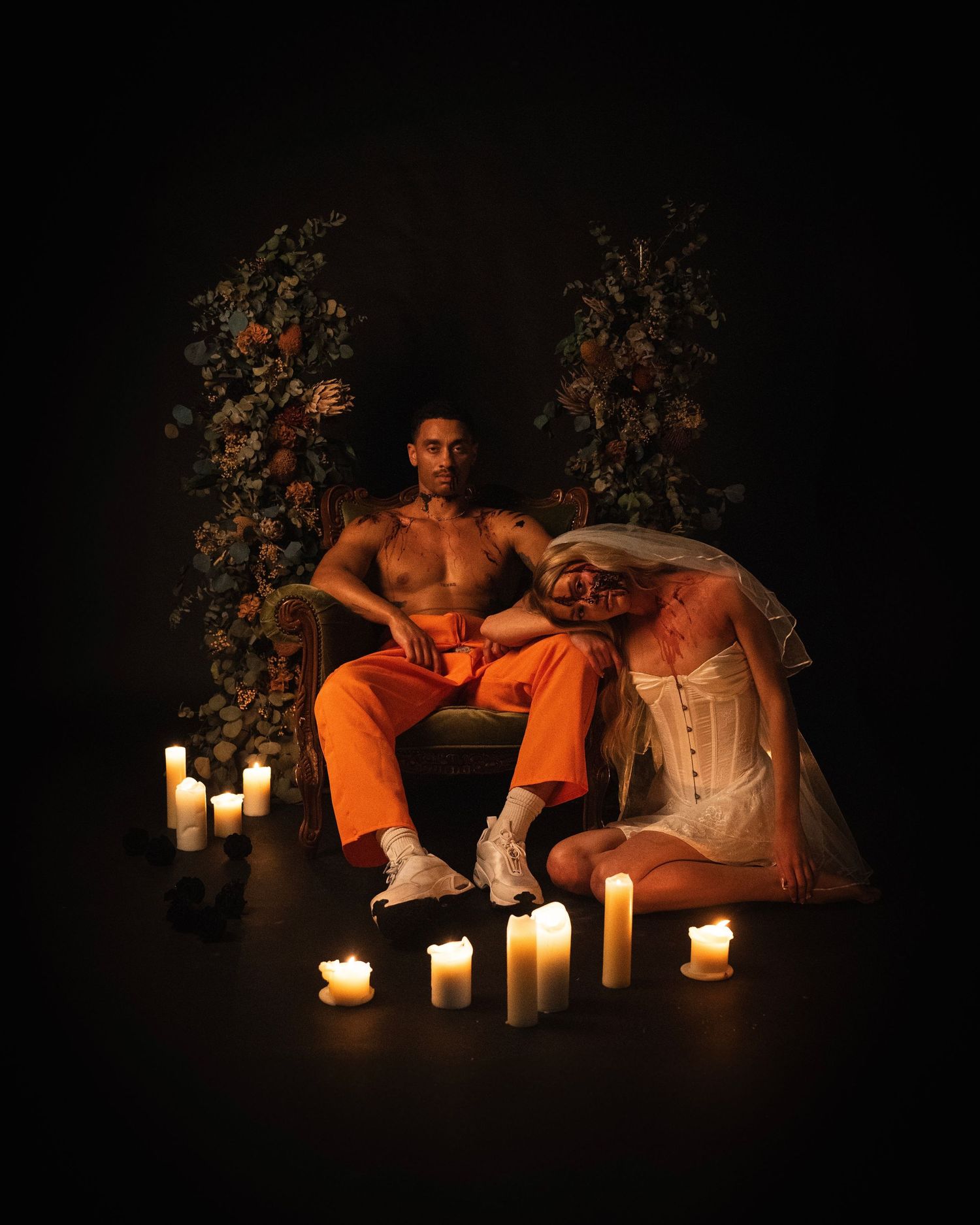 Two people in dark moody setting with glowing candles and floral arrangements create dramatic artistic portrait.