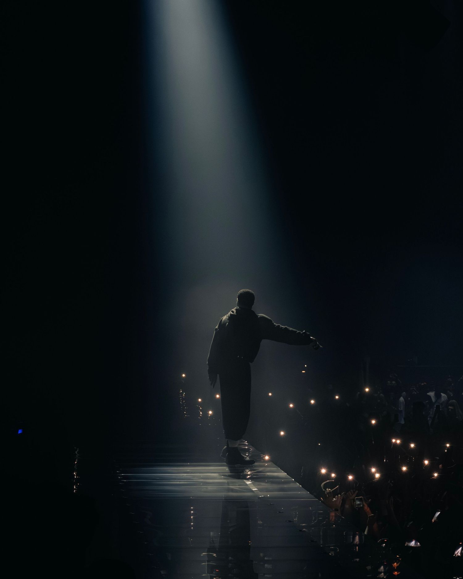 A silhouetted performer stands in a dramatic spotlight on stage while audience members hold up glowing lights in the darkness.