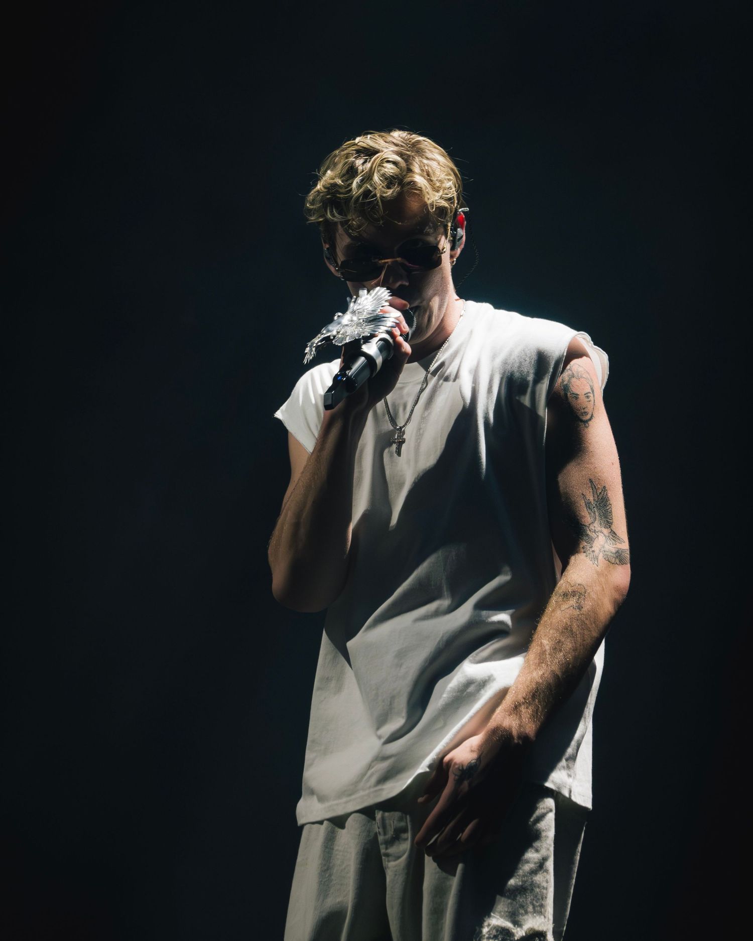 Performer in white sleeveless shirt holds microphone on dark stage under dramatic lighting during concert performance.