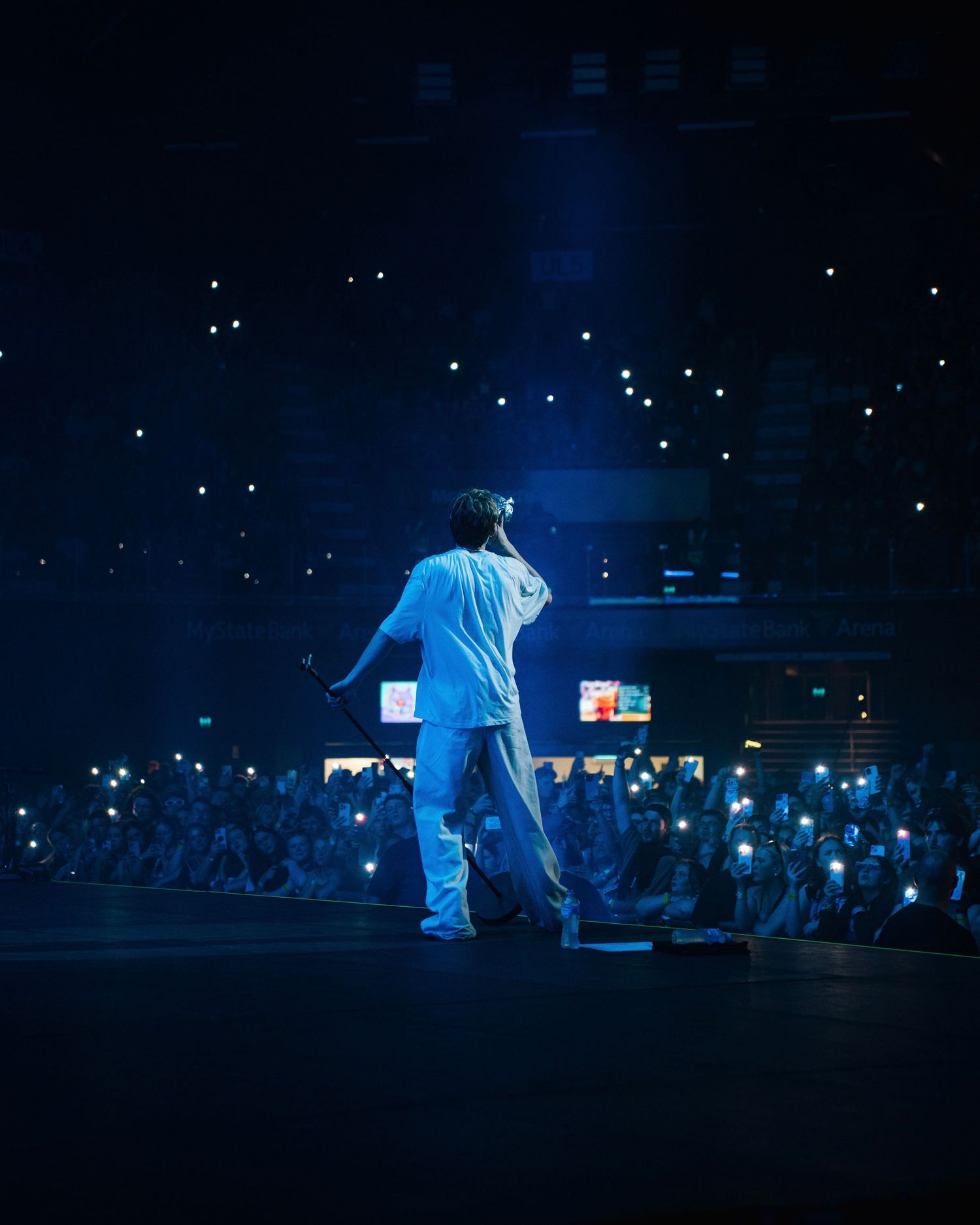 A performer in light blue clothing stands on a dark stage with glowing audience phone lights in the background.