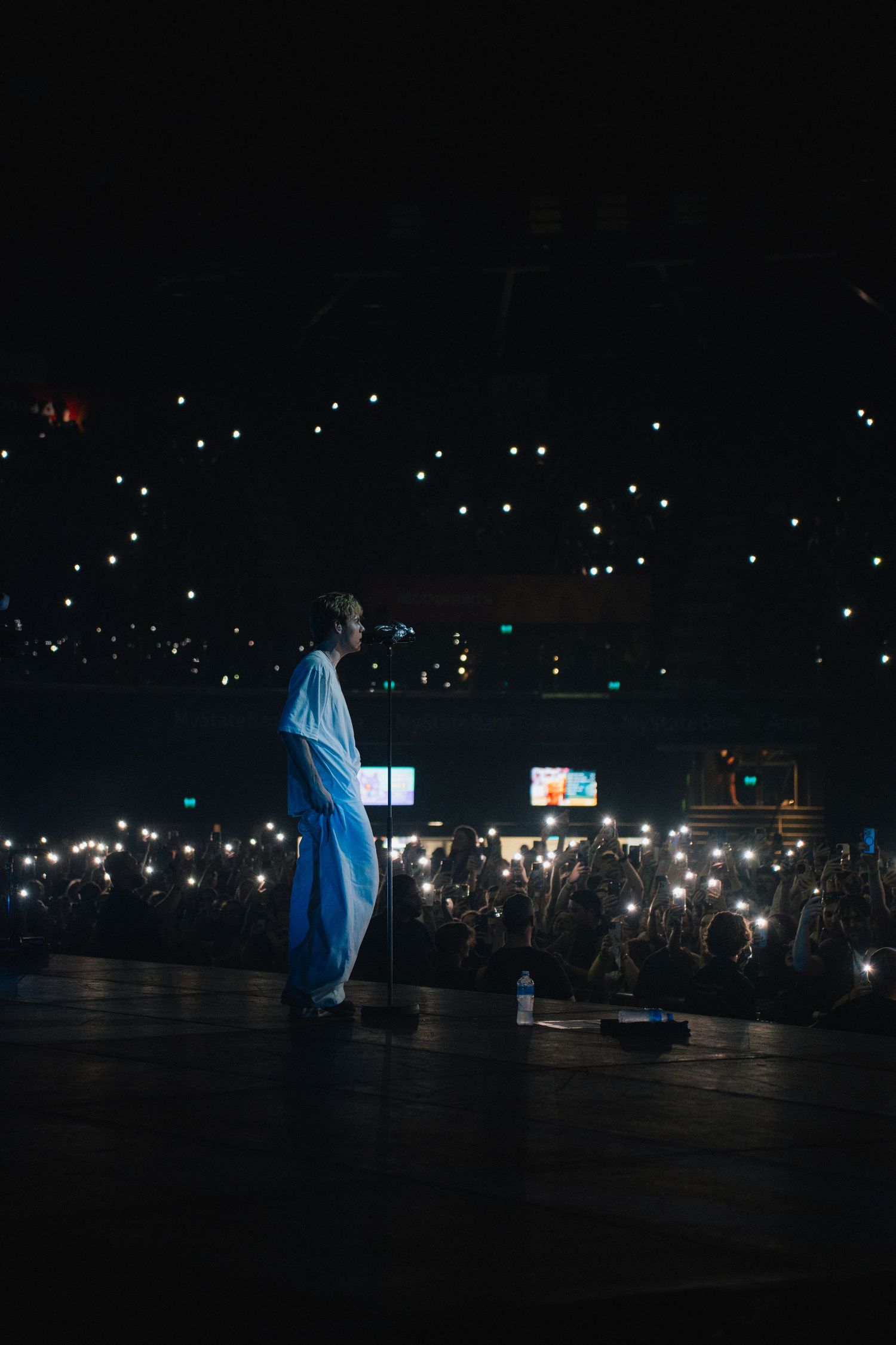 A silhouetted performer stands on a dark stage with a sea of glowing phone lights from the audience creating a magical atmosphere.