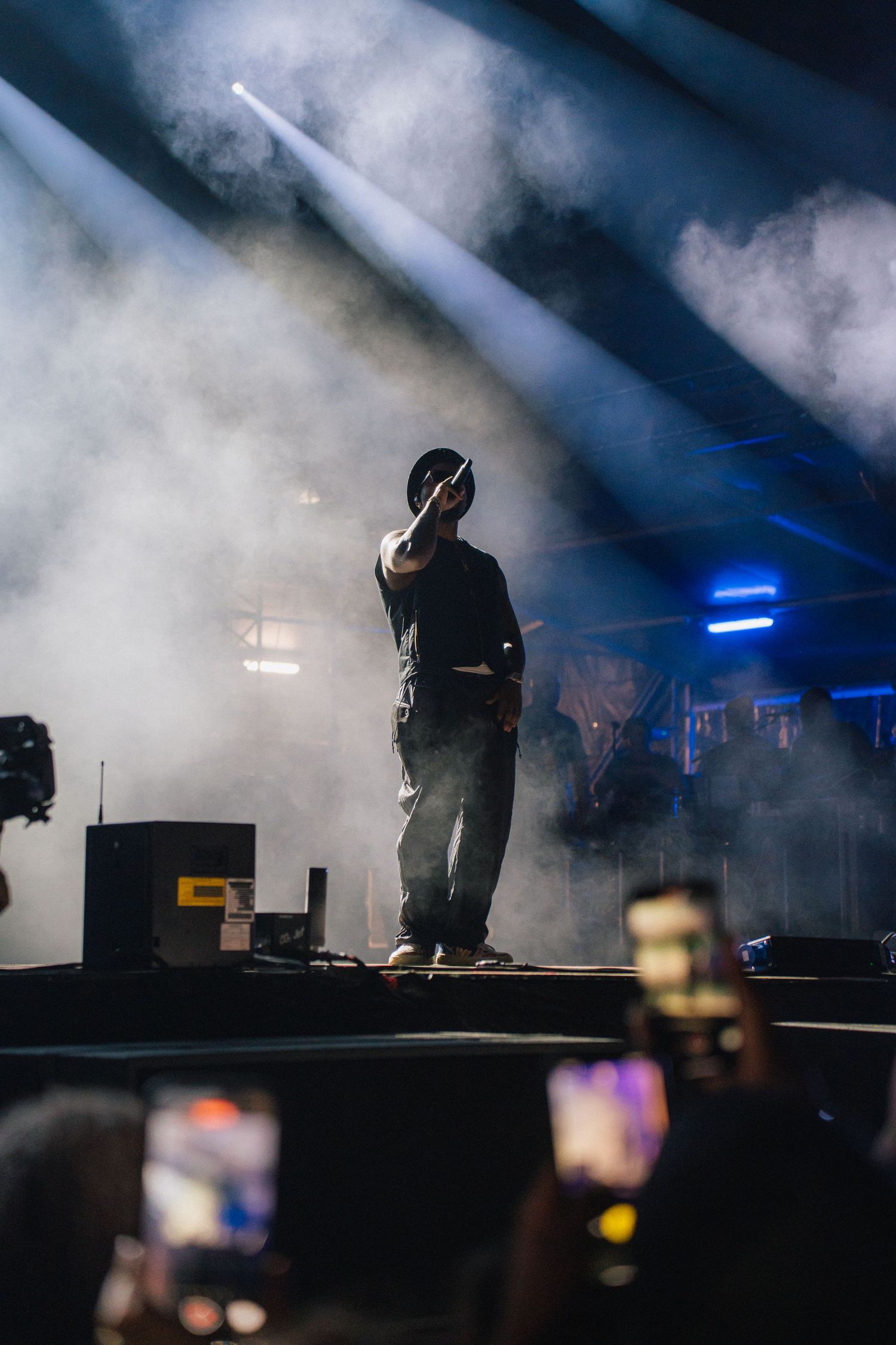 A performer stands on a misty stage in silhouette with blue lighting while audience members record on phones below.