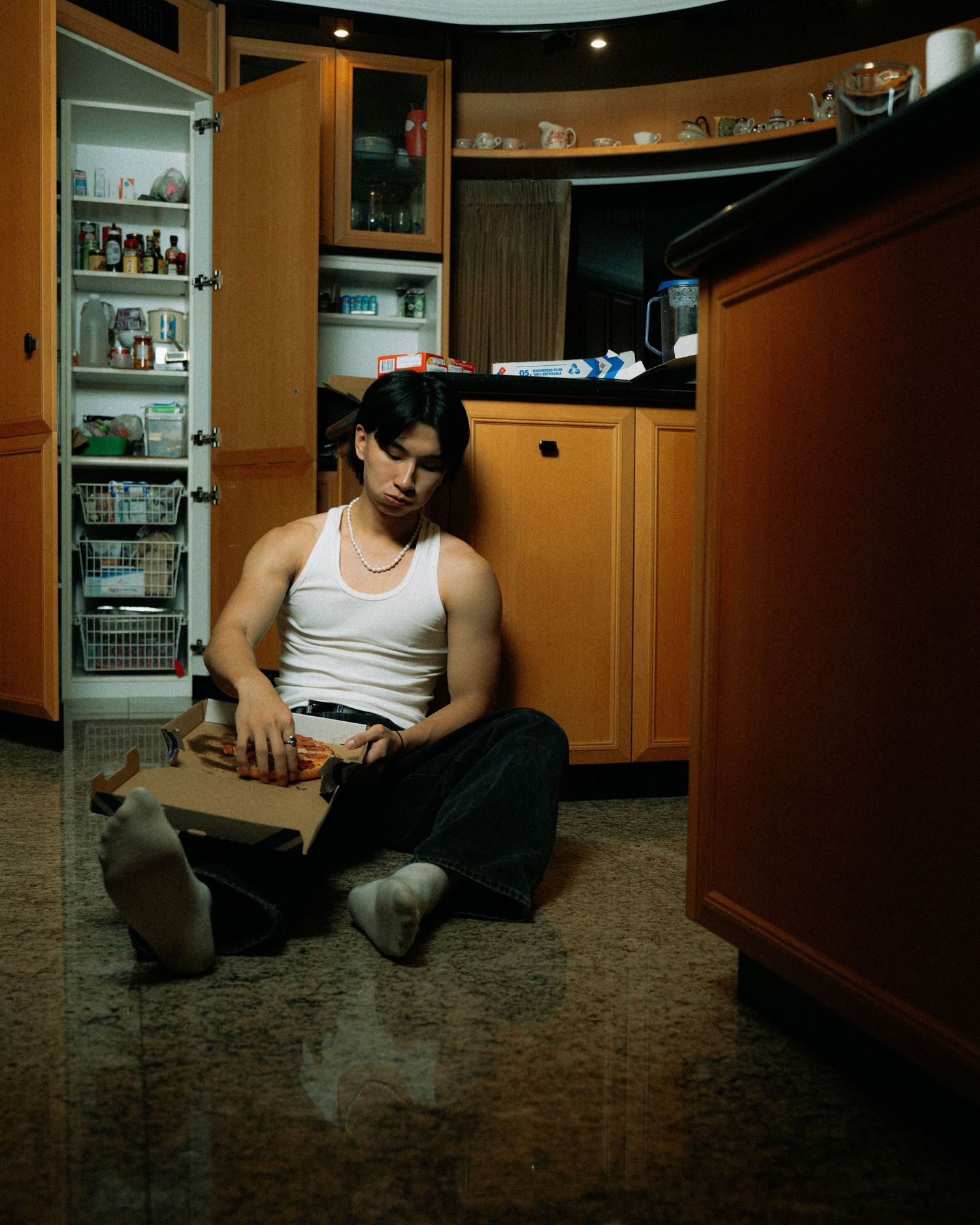 Someone sits on kitchen floor at night near wooden cabinets with illuminated storage shelves in background.