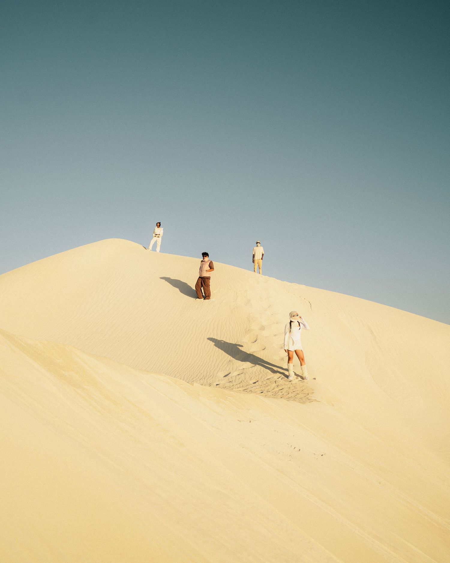 People sandboarding and playing on large desert sand dunes under blue sky.