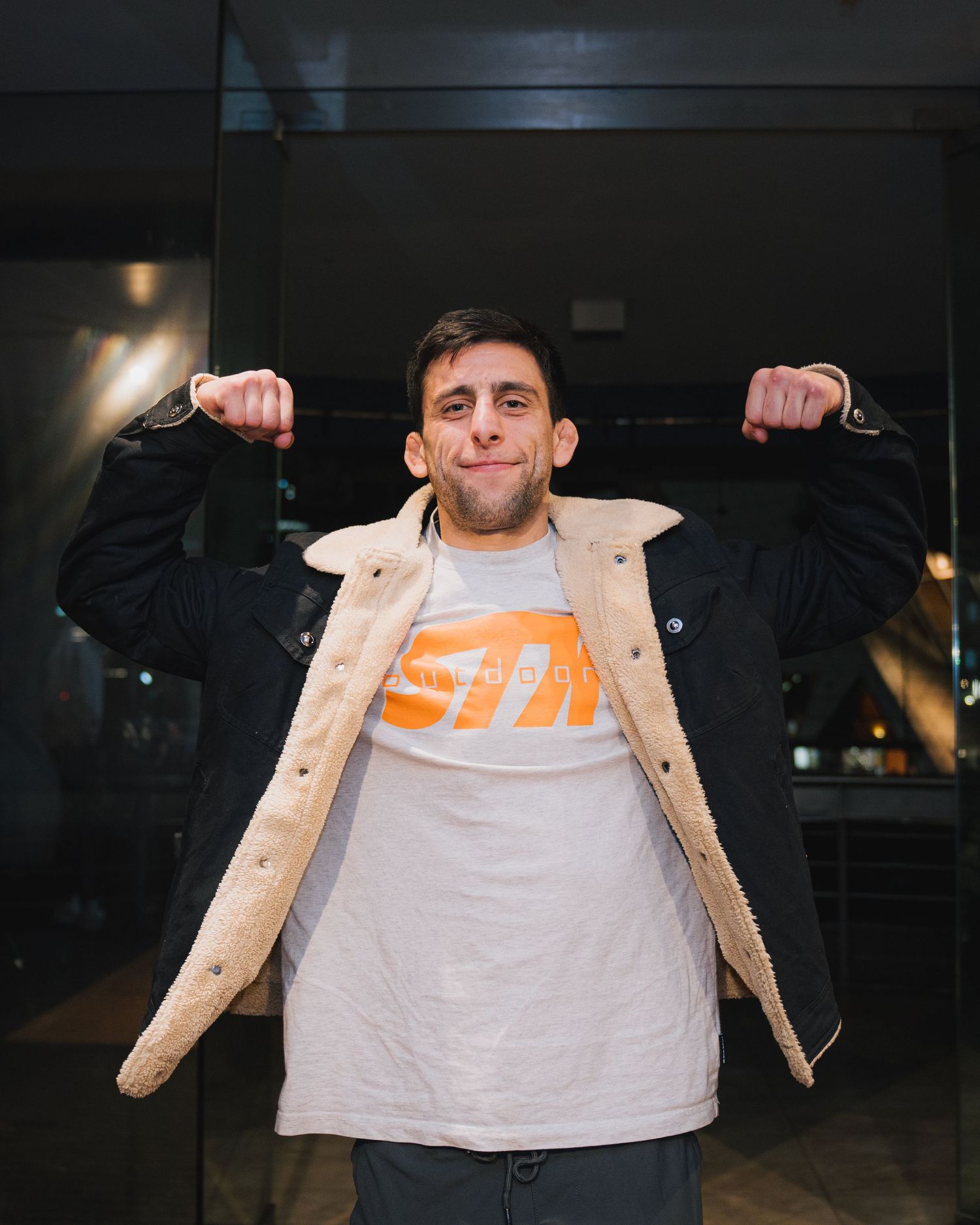 Person wearing tan jacket over orange graphic tee poses playfully indoors.