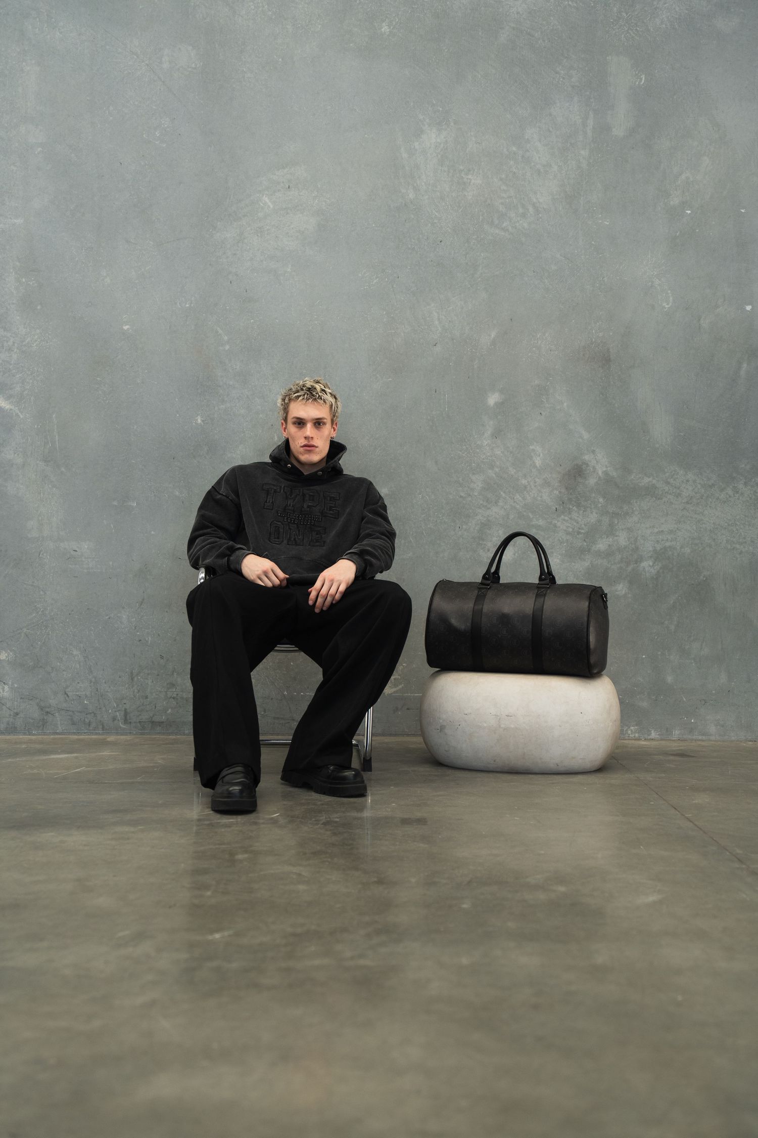 Fashion model in all-black outfit sits with duffle bag on concrete platform against grey wall.