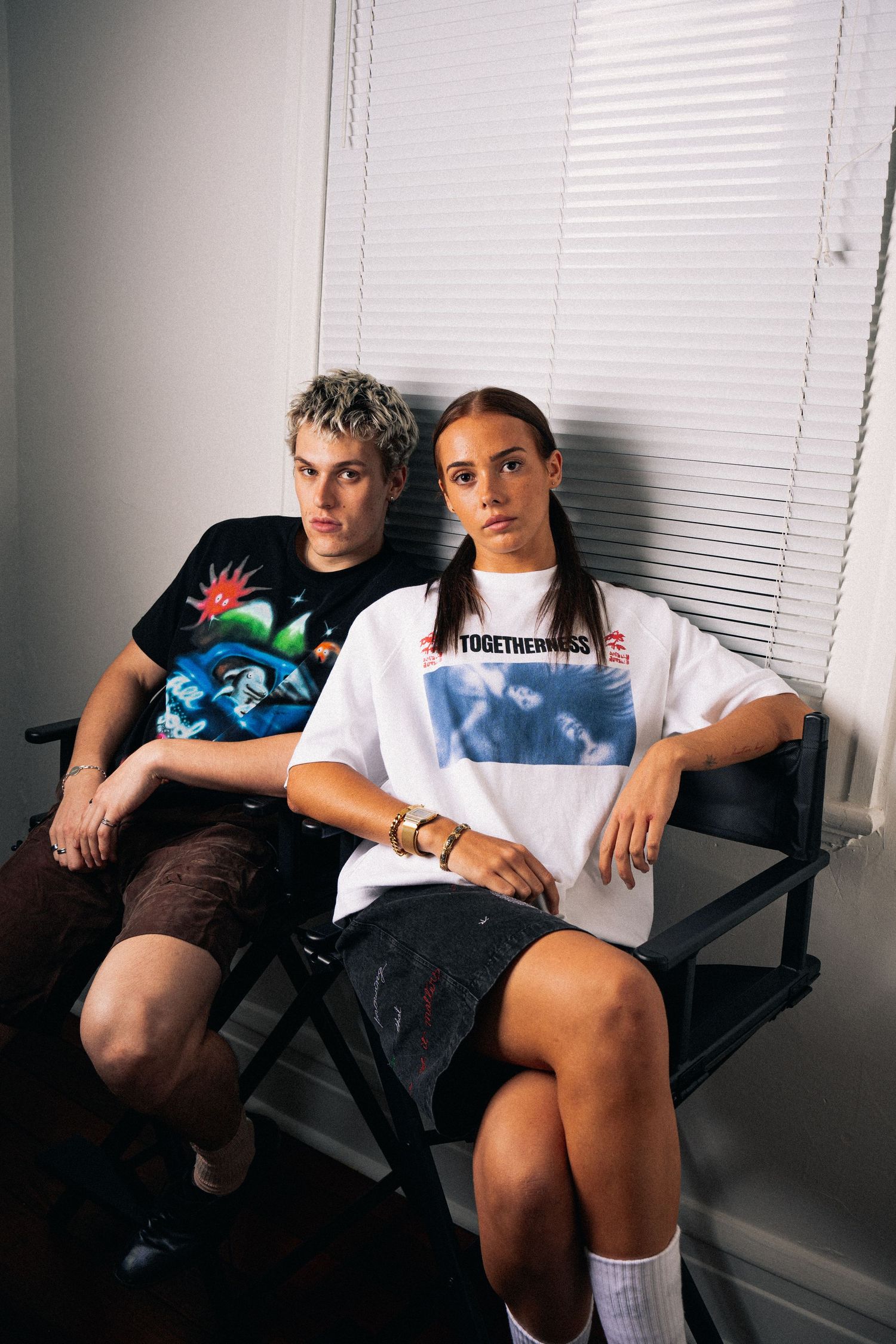 Two people sitting casually on director's chairs modeling casual streetwear.