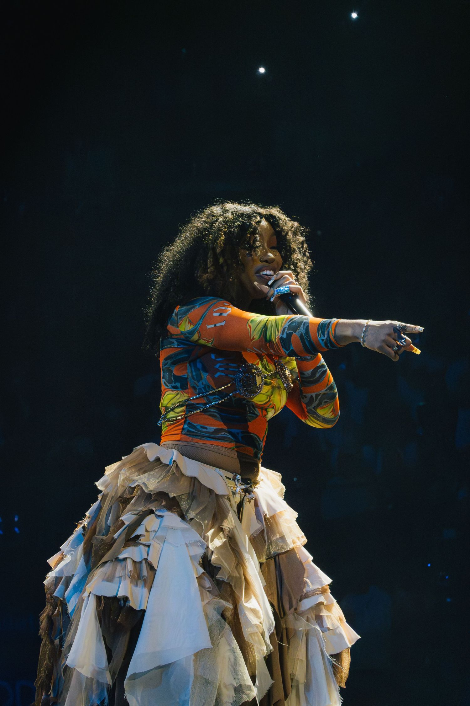 Performer in colorful patterned top and layered white skirt performs energetically on darkened stage with spotlights above.