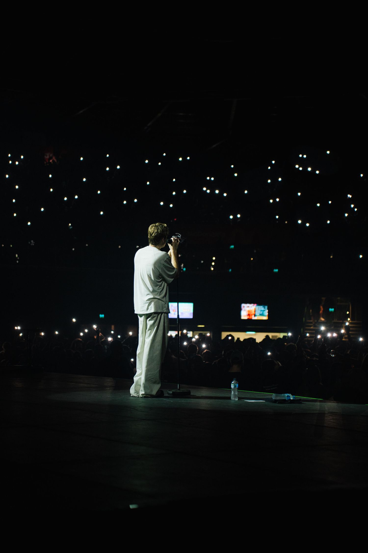 A performer stands silhouetted on a dark stage with glowing phone lights scattered throughout the audience in the background.