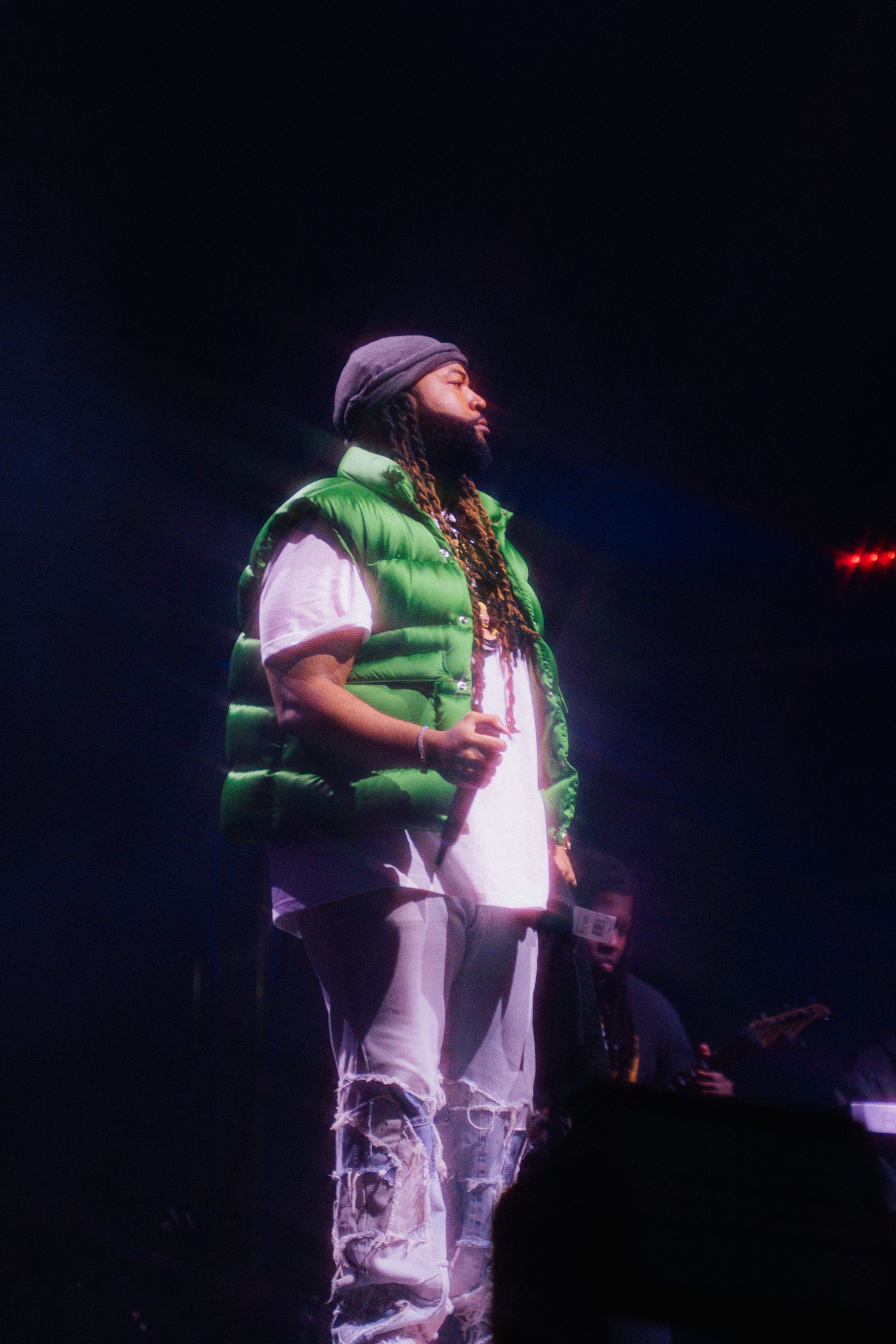 Performer on stage wearing a green vest, white pants and purple beanie during a concert performance in dark lighting.