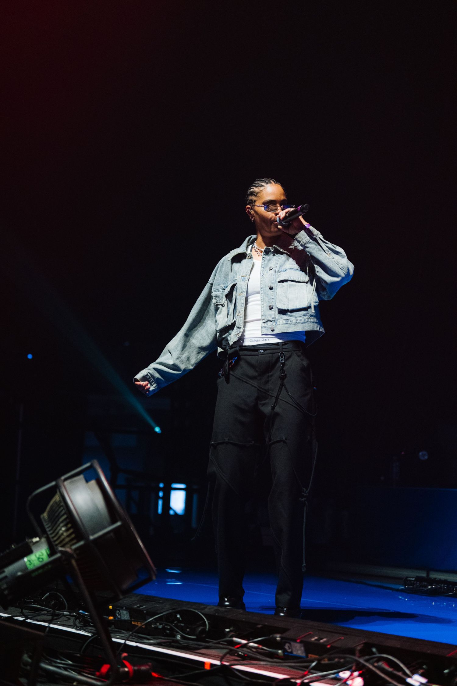 Performer in light denim jacket and black pants sings on darkened stage with dramatic lighting and equipment setup.