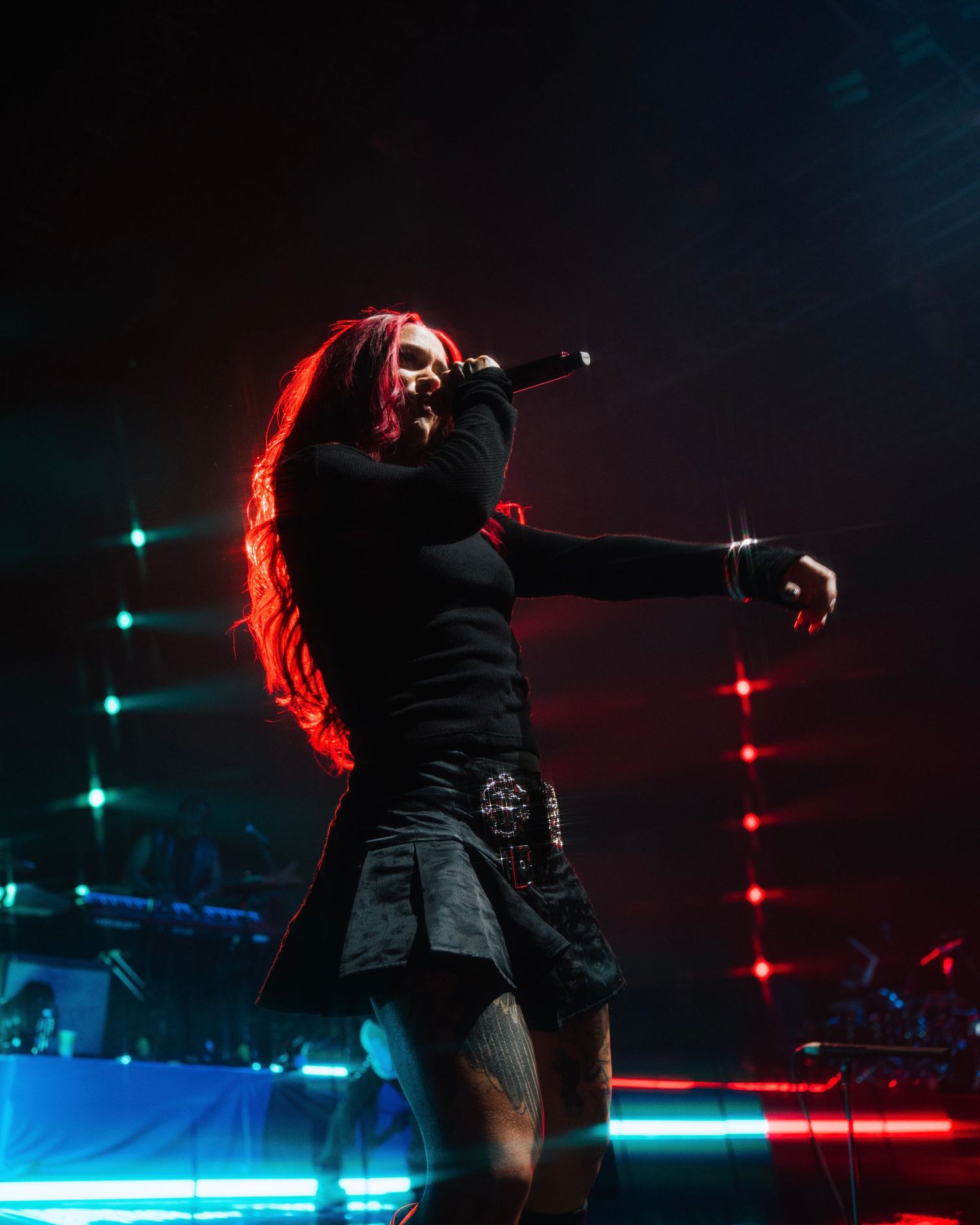 A performer in black clothing under red and blue stage lighting reaches out with dramatic gestures during a concert performance.