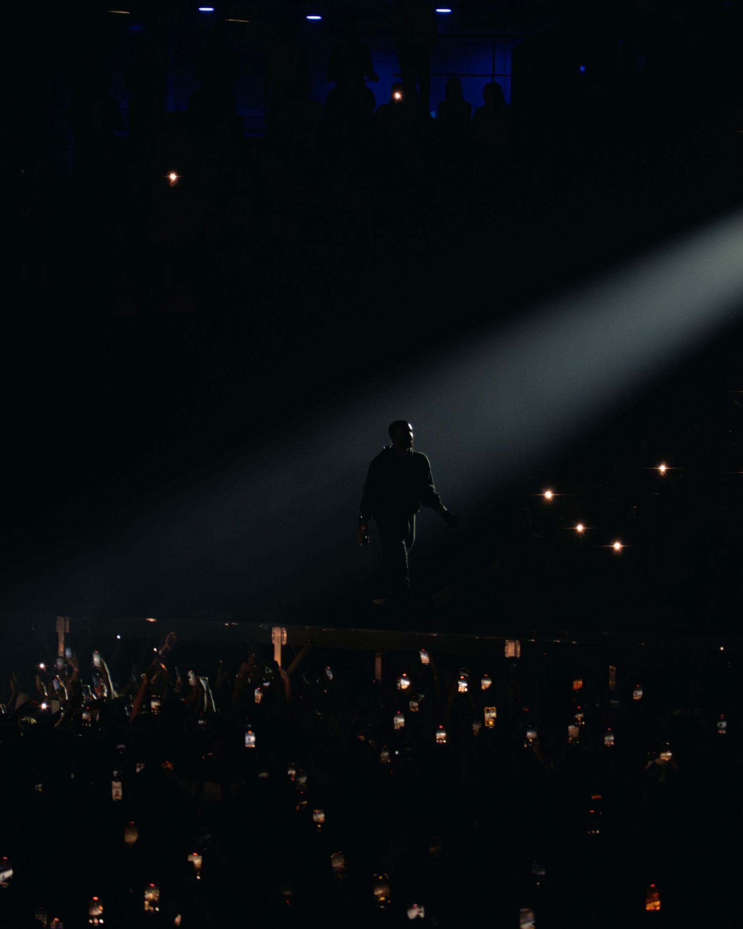 A silhouetted performer stands in a spotlight on stage with glowing lights from the audience below in a dark venue.