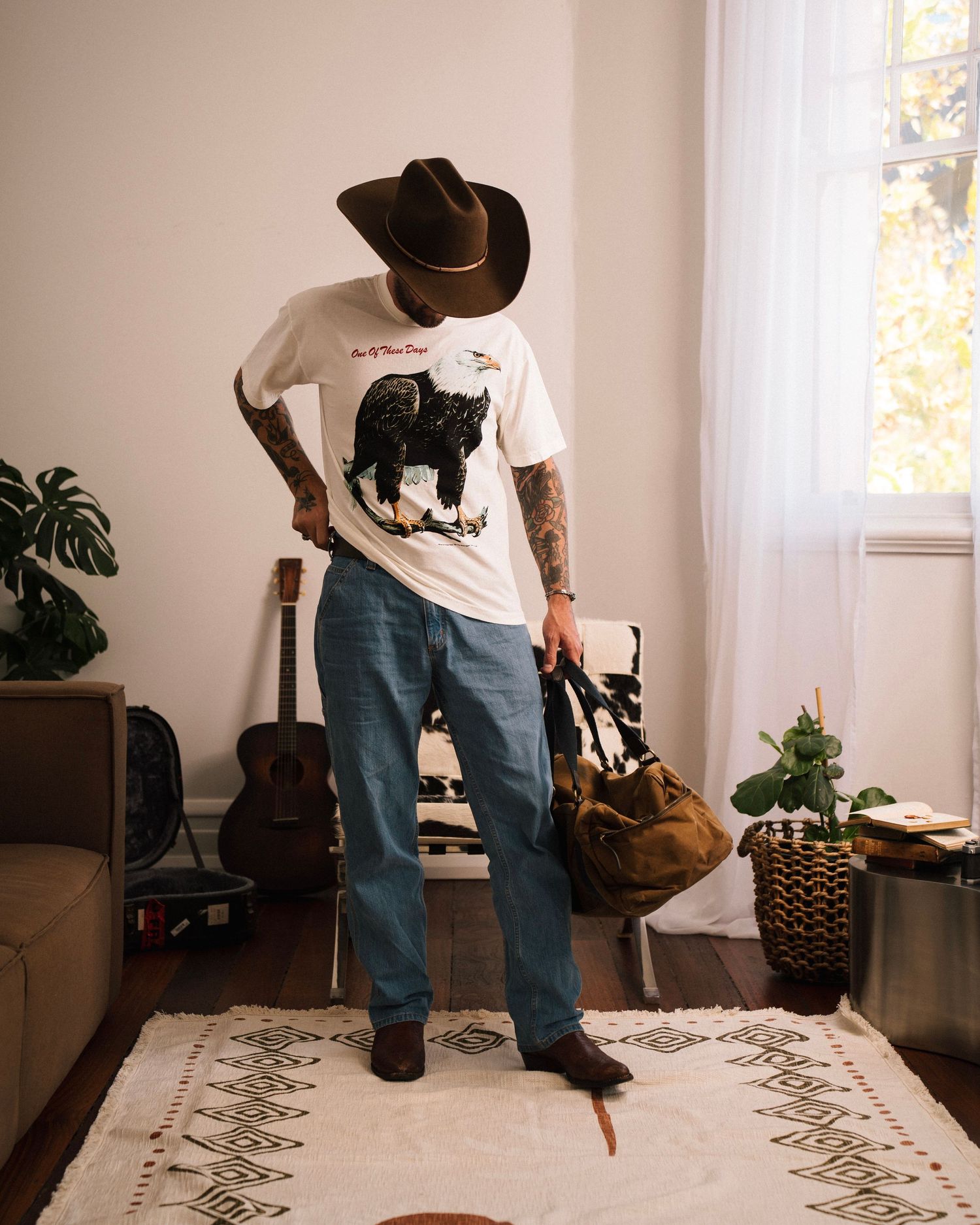 Person in cowboy hat and graphic t-shirt stands on patterned rug with guitar in background.