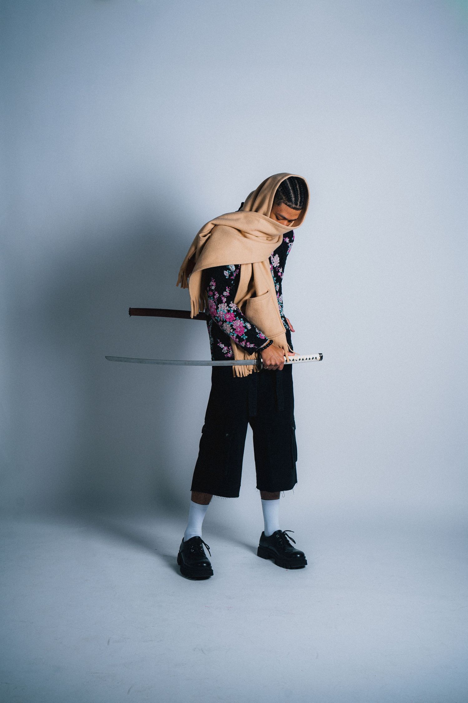 Fashion model poses with katana sword wearing floral print top and black pants against grey backdrop.