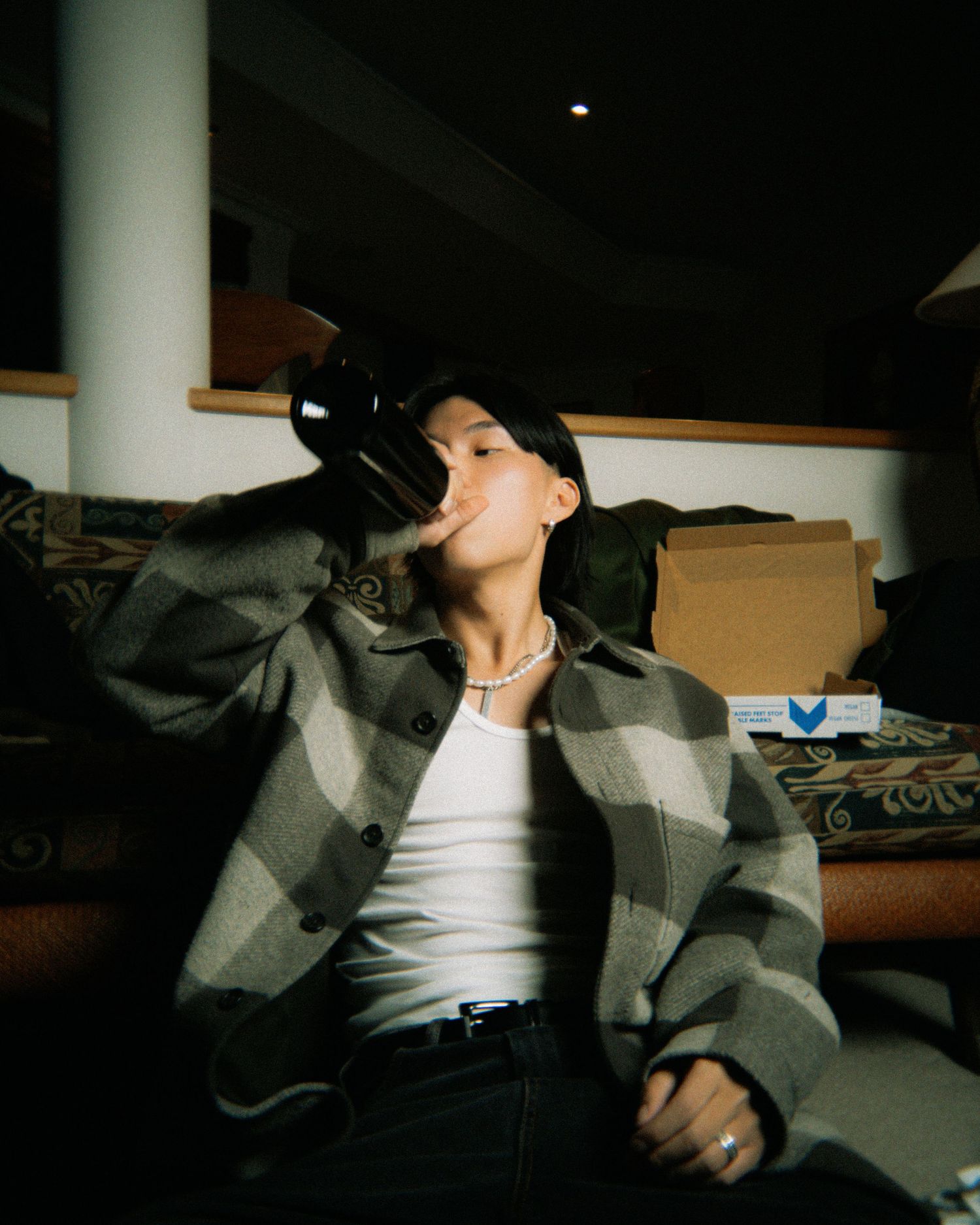 Person wearing a plaid jacket and white tank top sits casually while looking up in a moody indoor setting.