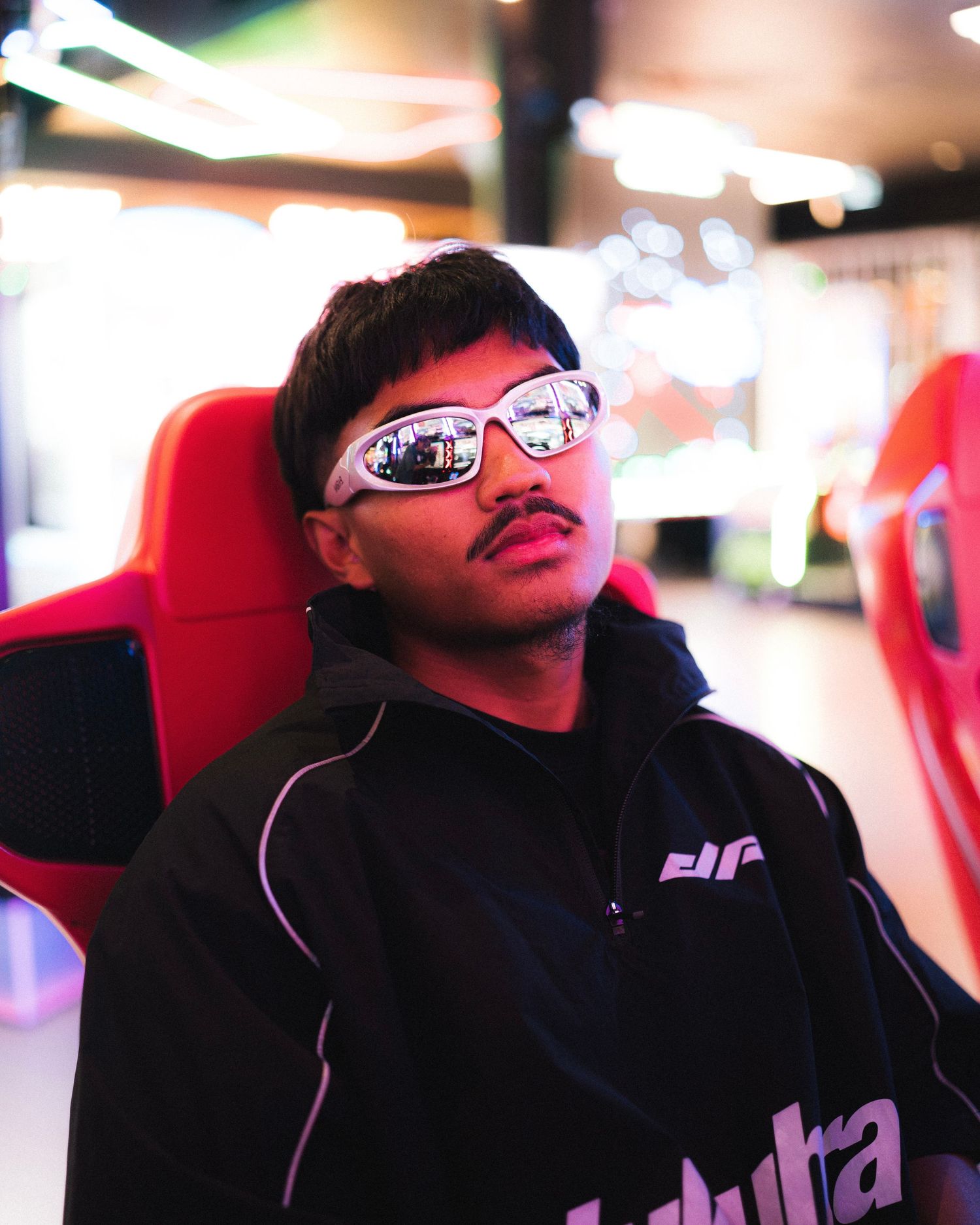 Person with mirrored sunglasses wearing a dark colored hoodie sits in a red gaming chair under indoor lighting.