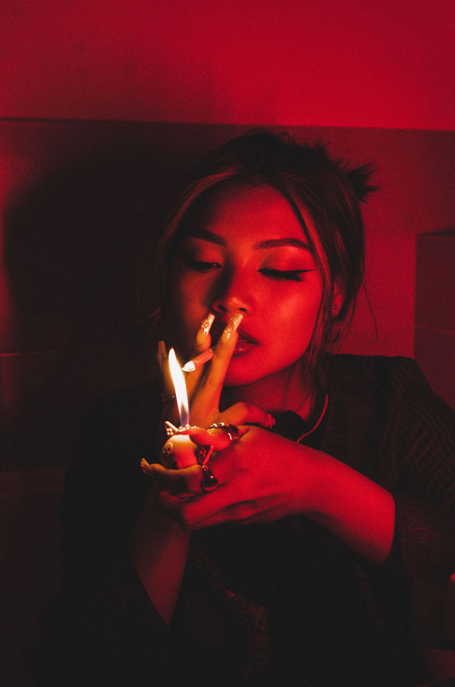 Portrait lit by dramatic red lighting shows figure lighting multiple cigarettes in dark moody atmosphere.