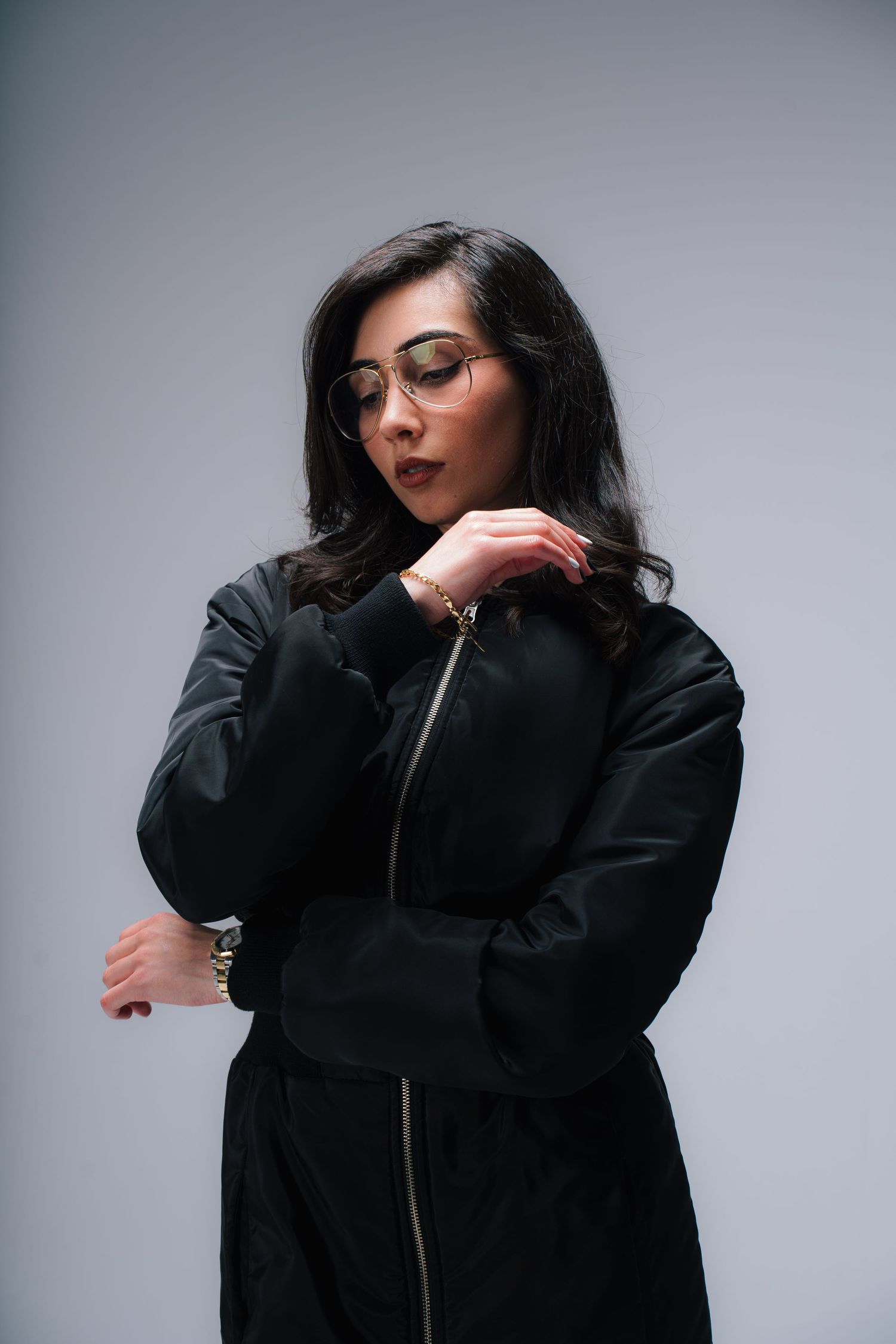 A figure wearing glasses and a black winter coat poses against a gray background with a contemplative expression.