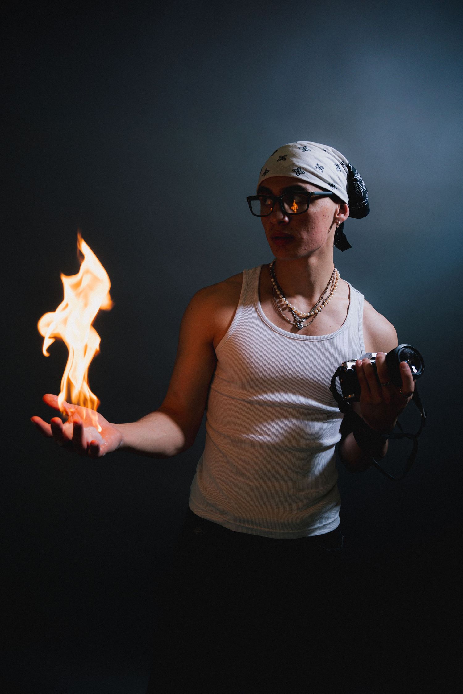 A person in a white tank top, headphones and glasses performs fire juggling on a dark background.