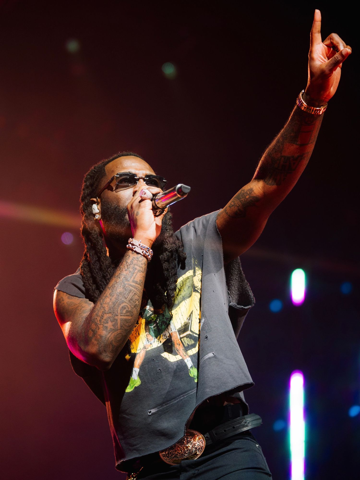 A performer in a black t-shirt sings into a microphone with one arm raised on a colorfully lit concert stage.
