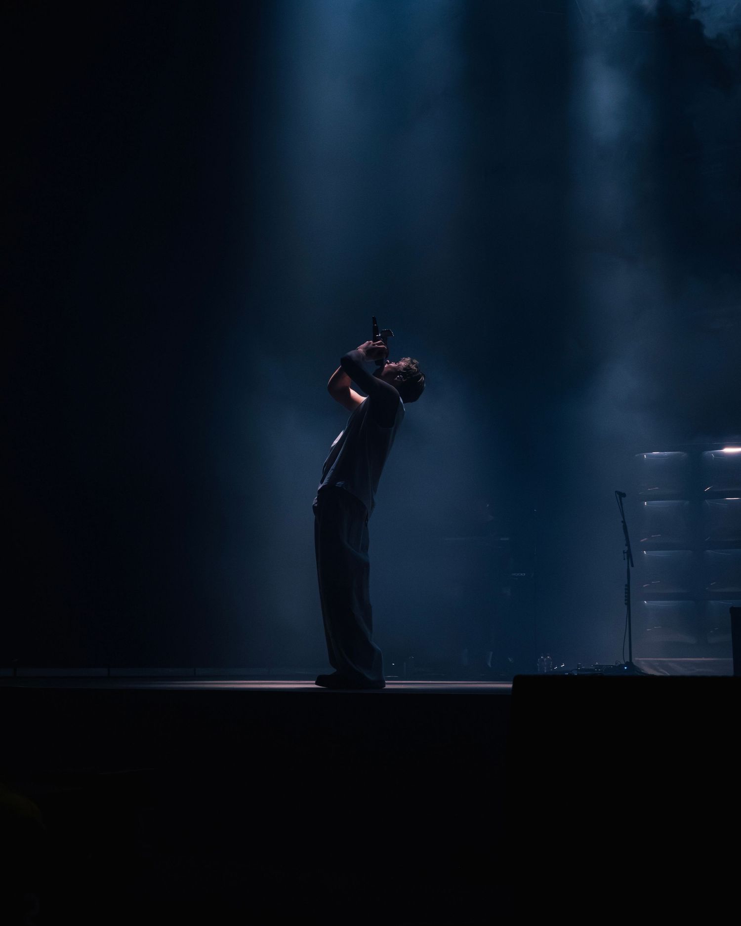 A performer stands in silhouette on a dark stage with dramatic blue spotlights creating an atmospheric scene.