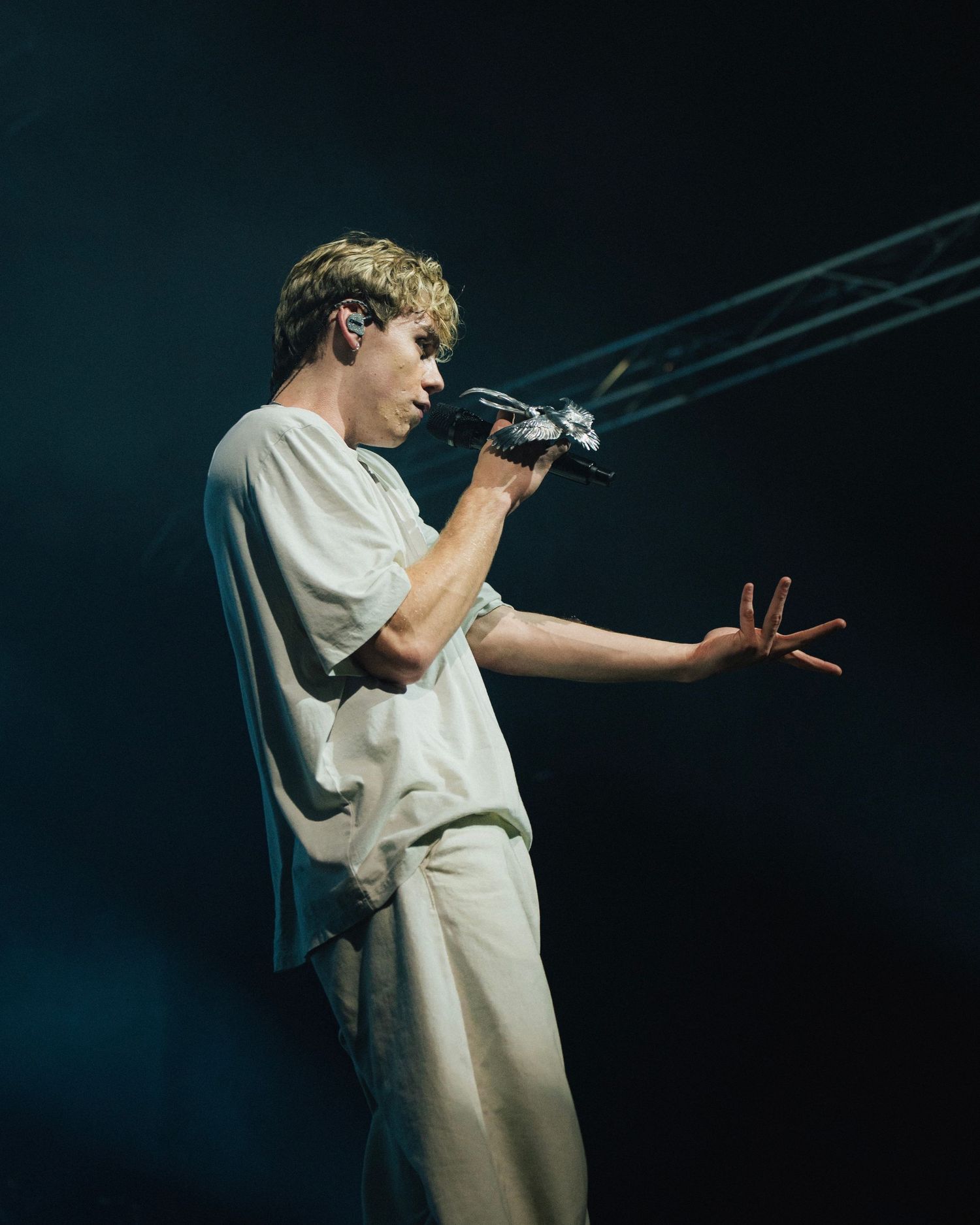 A performer in white attire gestures expressively while singing into a microphone on a dimly lit stage.
