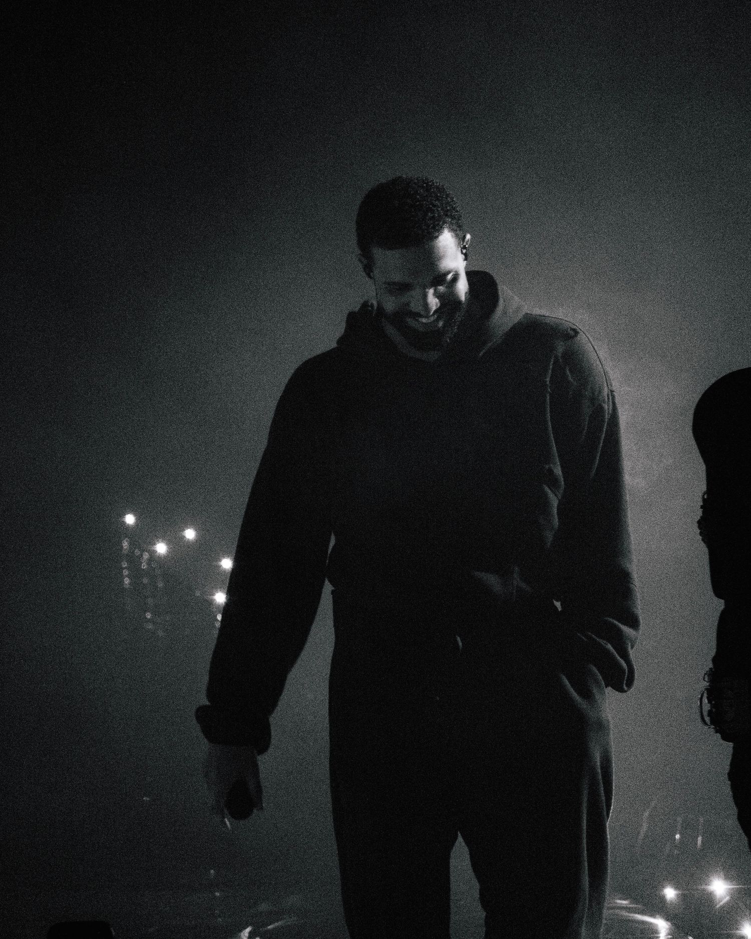 Atmospheric black and white photo of a person in dark clothing standing in dim lighting with bokeh lights in background.