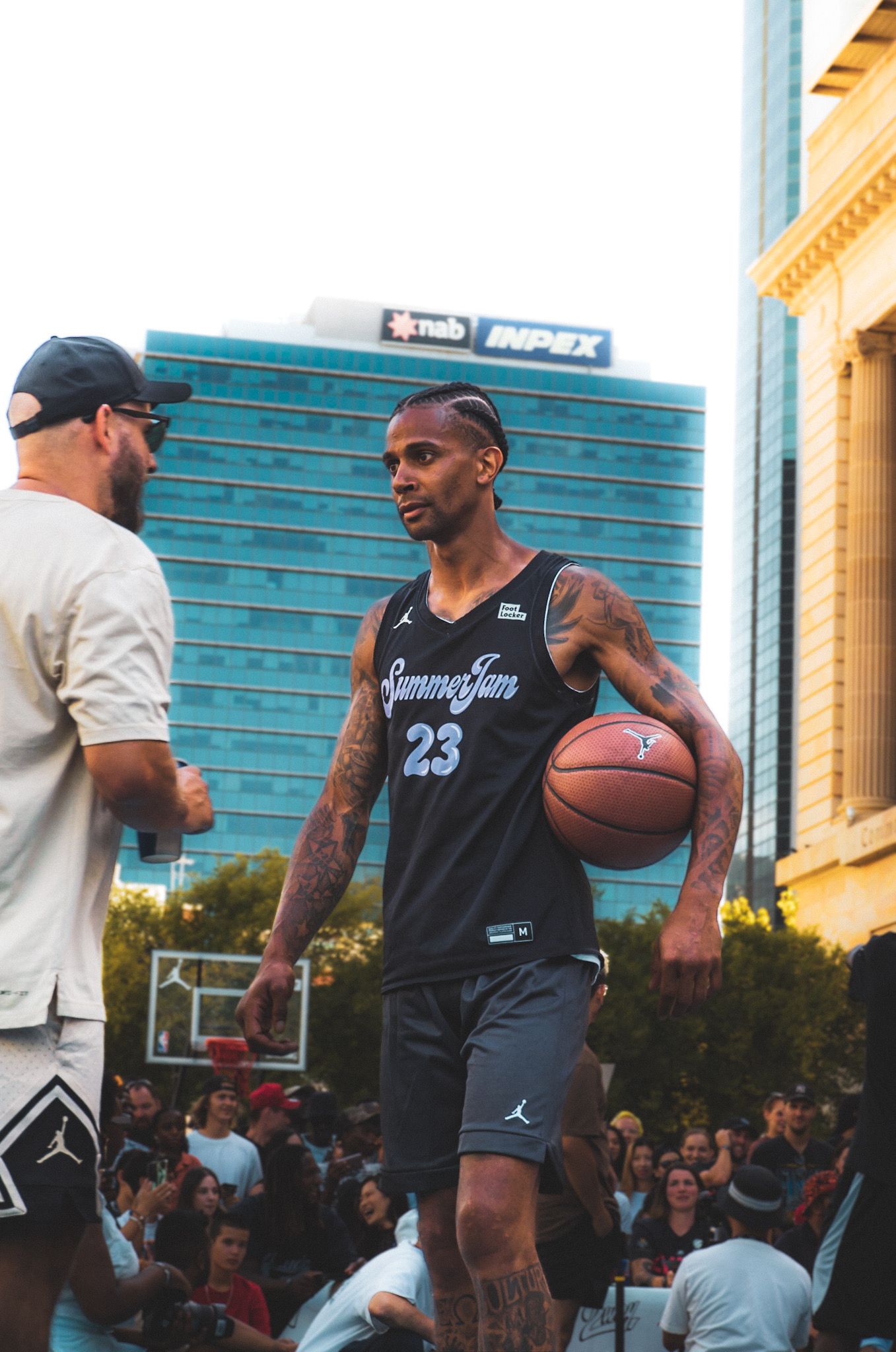 Basketball player wearing number 23 jersey holds ball during outdoor game with urban backdrop.