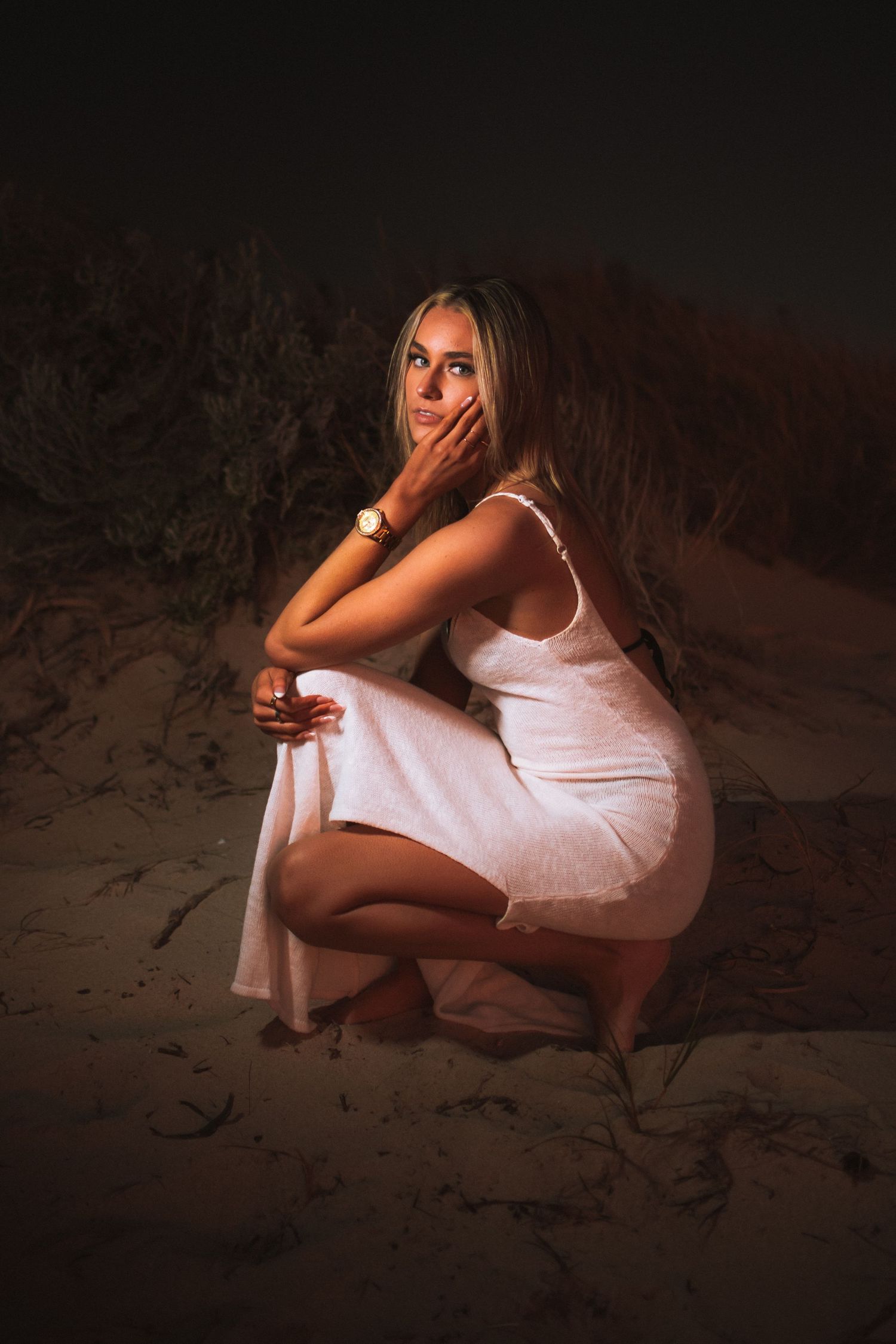 A woman in a white dress poses crouched on sand in moody lighting.