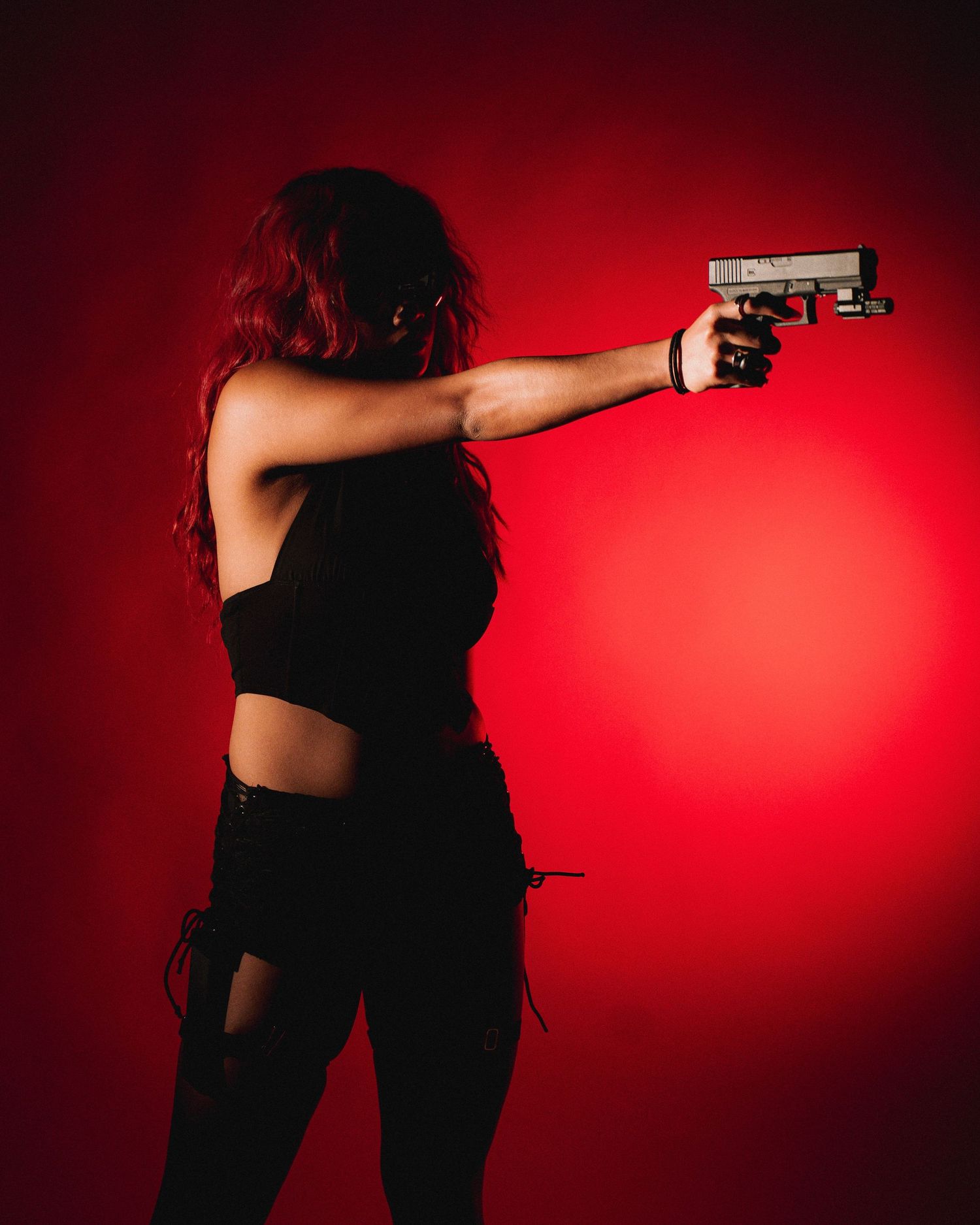 Figure in black outfit aims pistol against dramatic red background lighting in moody silhouetted pose.