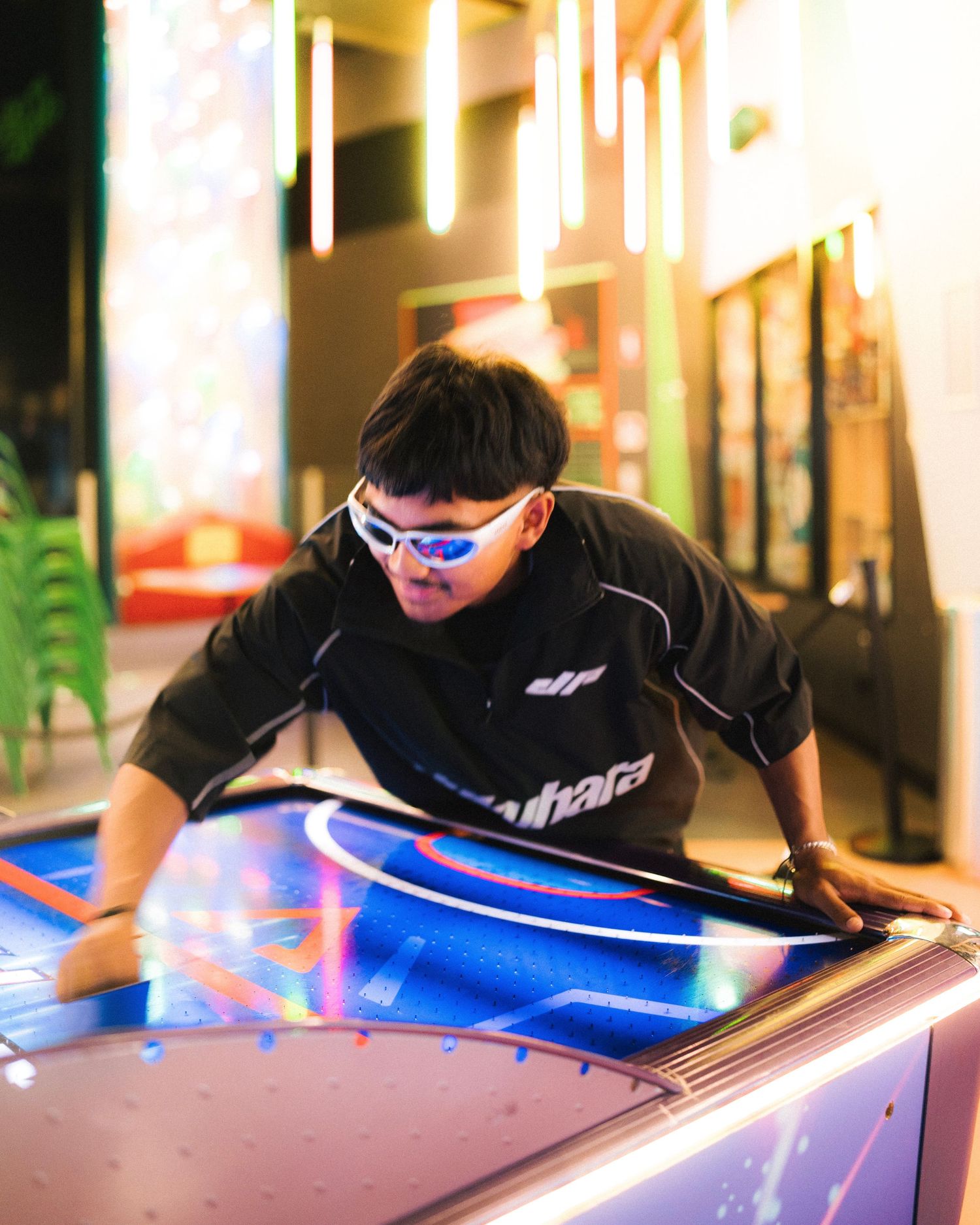Player in black shirt and sunglasses leans over an illuminated arcade game table in a modern entertainment venue.