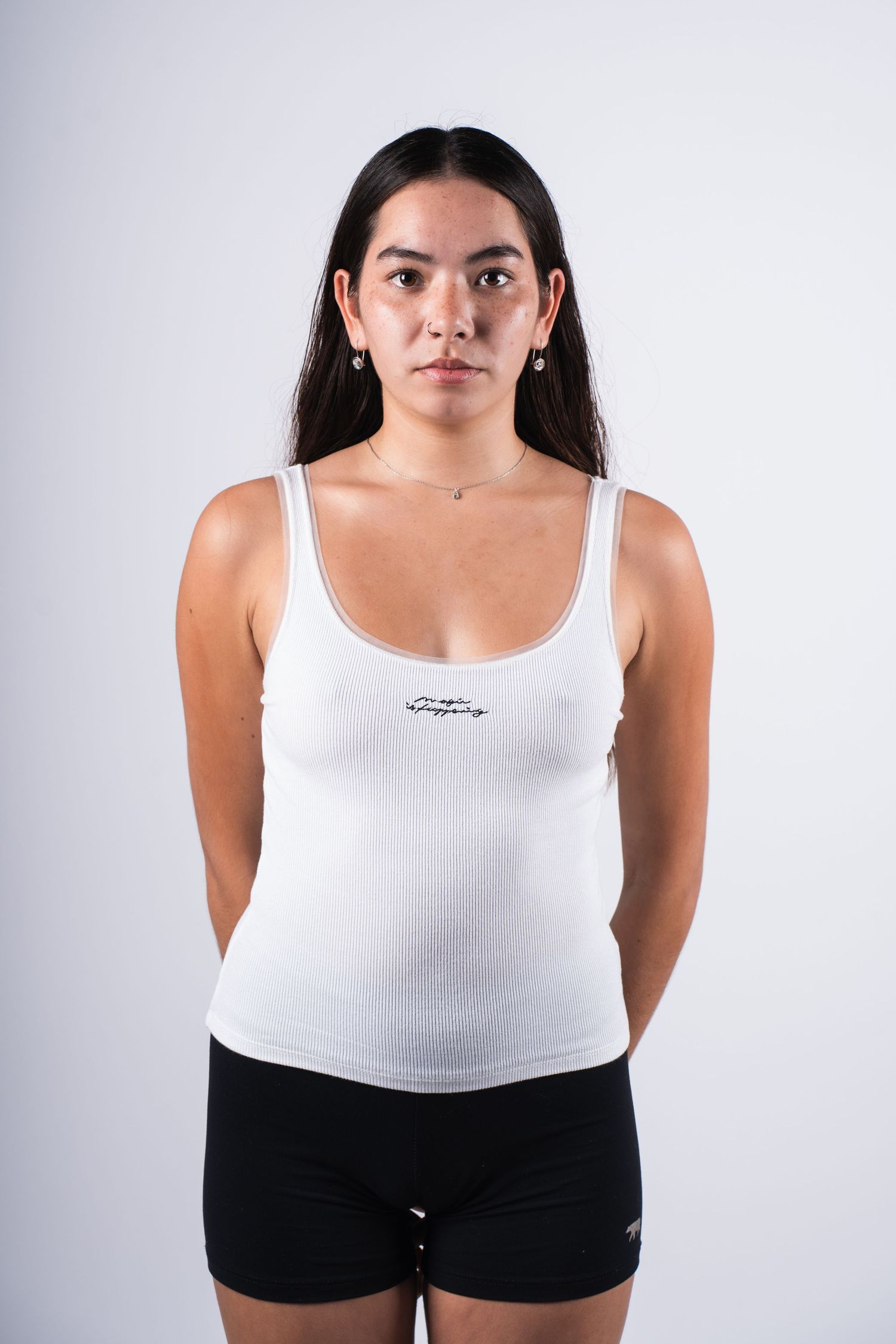 Person in athletic attire wearing a white tank top and black shorts stands against a grey background in a studio setting.