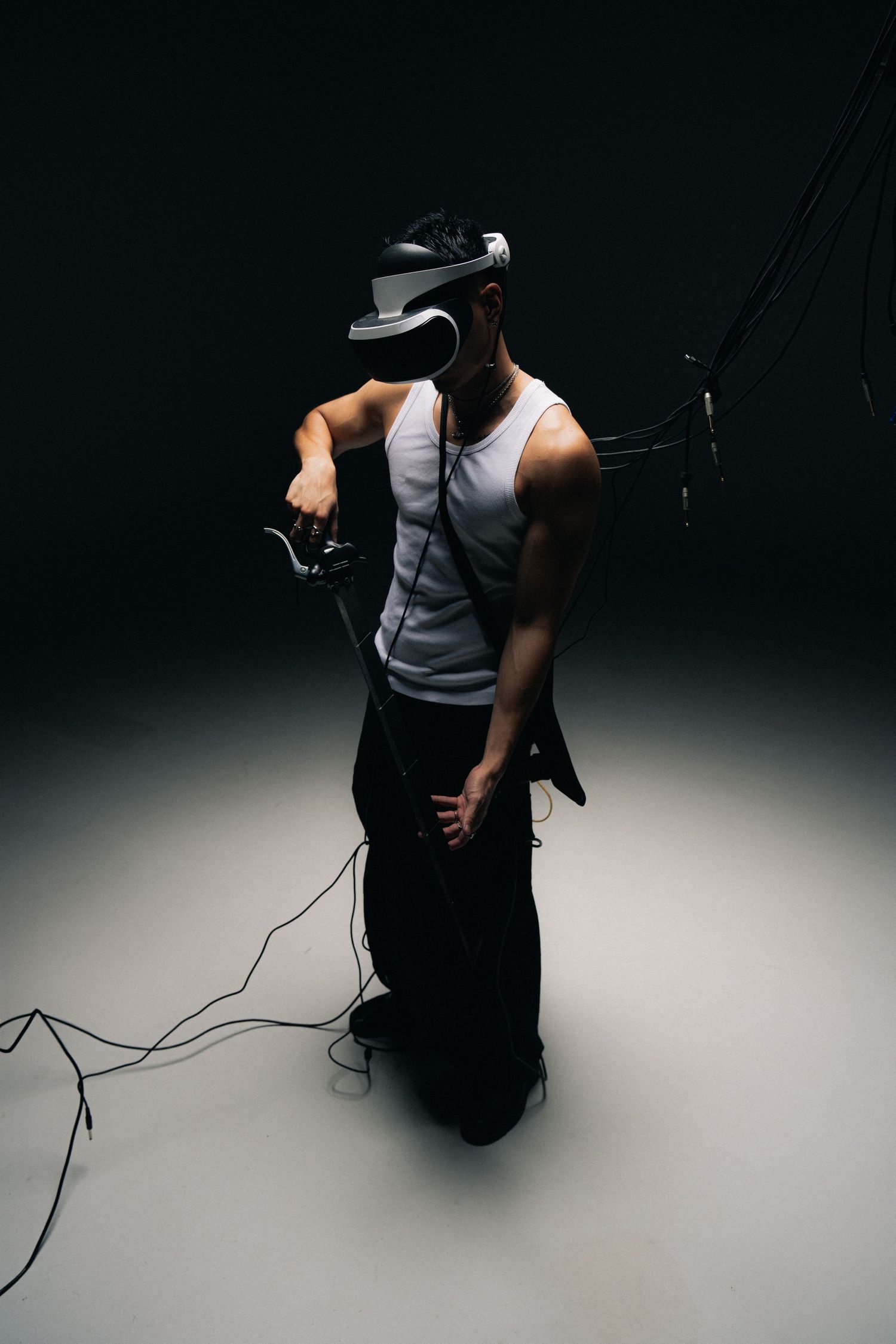 Person wearing VR headset and white tank top standing against dark background with cables attached to equipment.