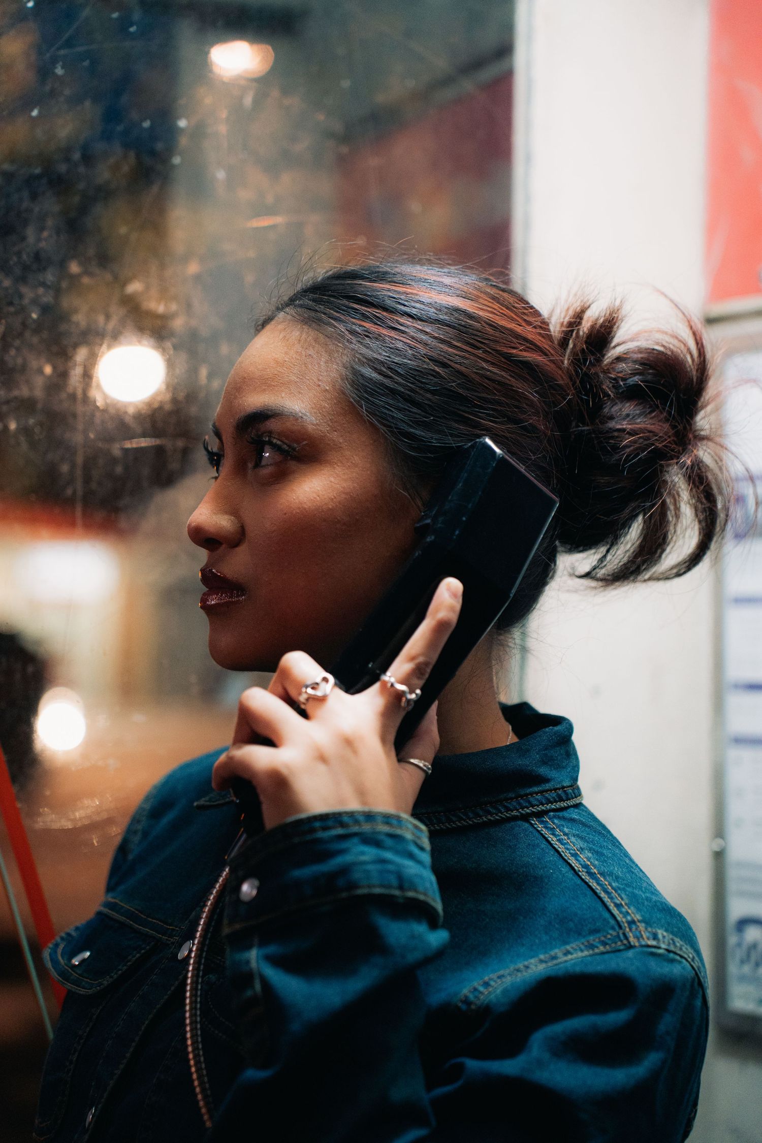Profile view of someone in a denim jacket talking on a mobile phone with city lights in the background.