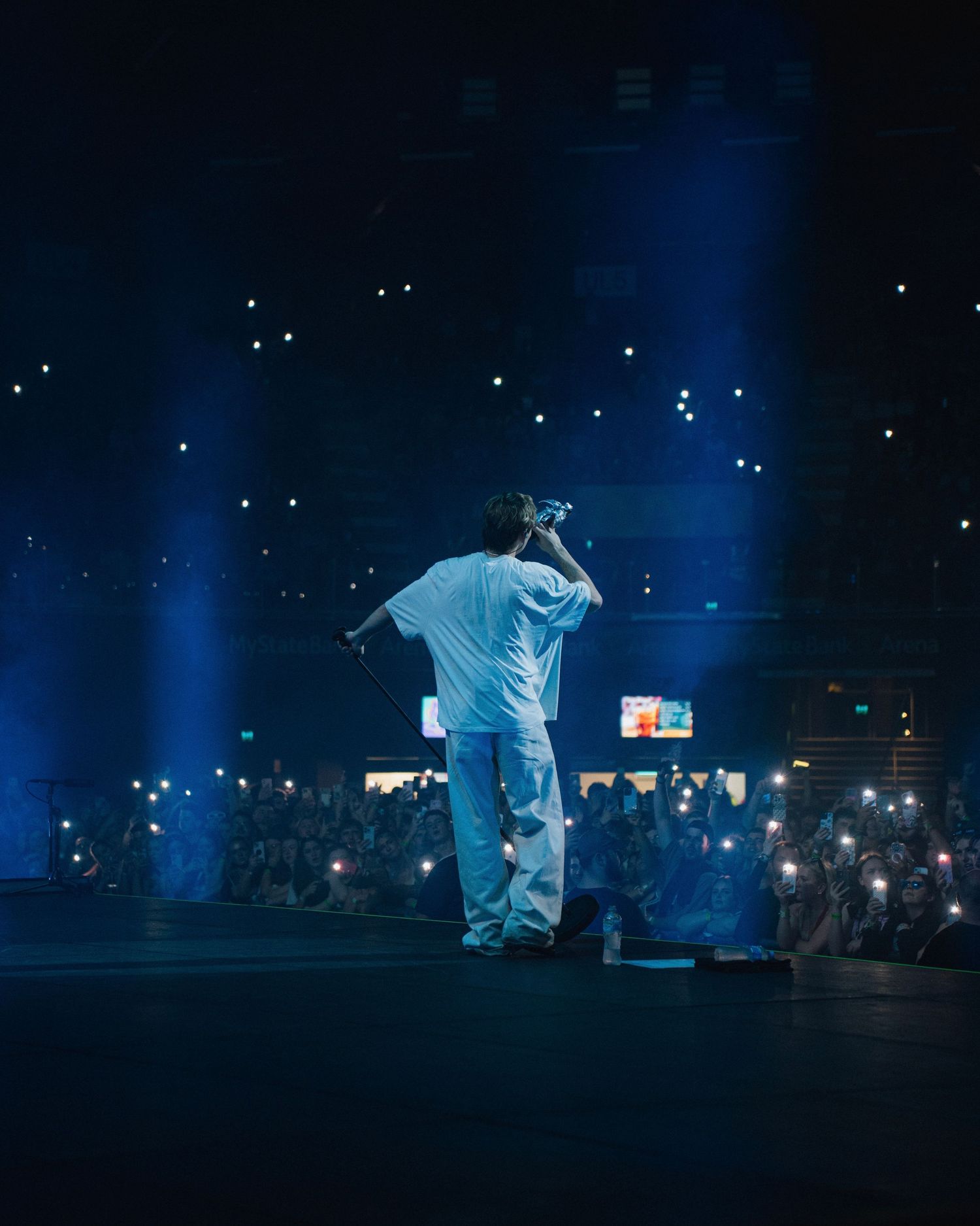 A performer in white clothing stands on a dark stage with blue lighting and glowing phone screens in the audience.