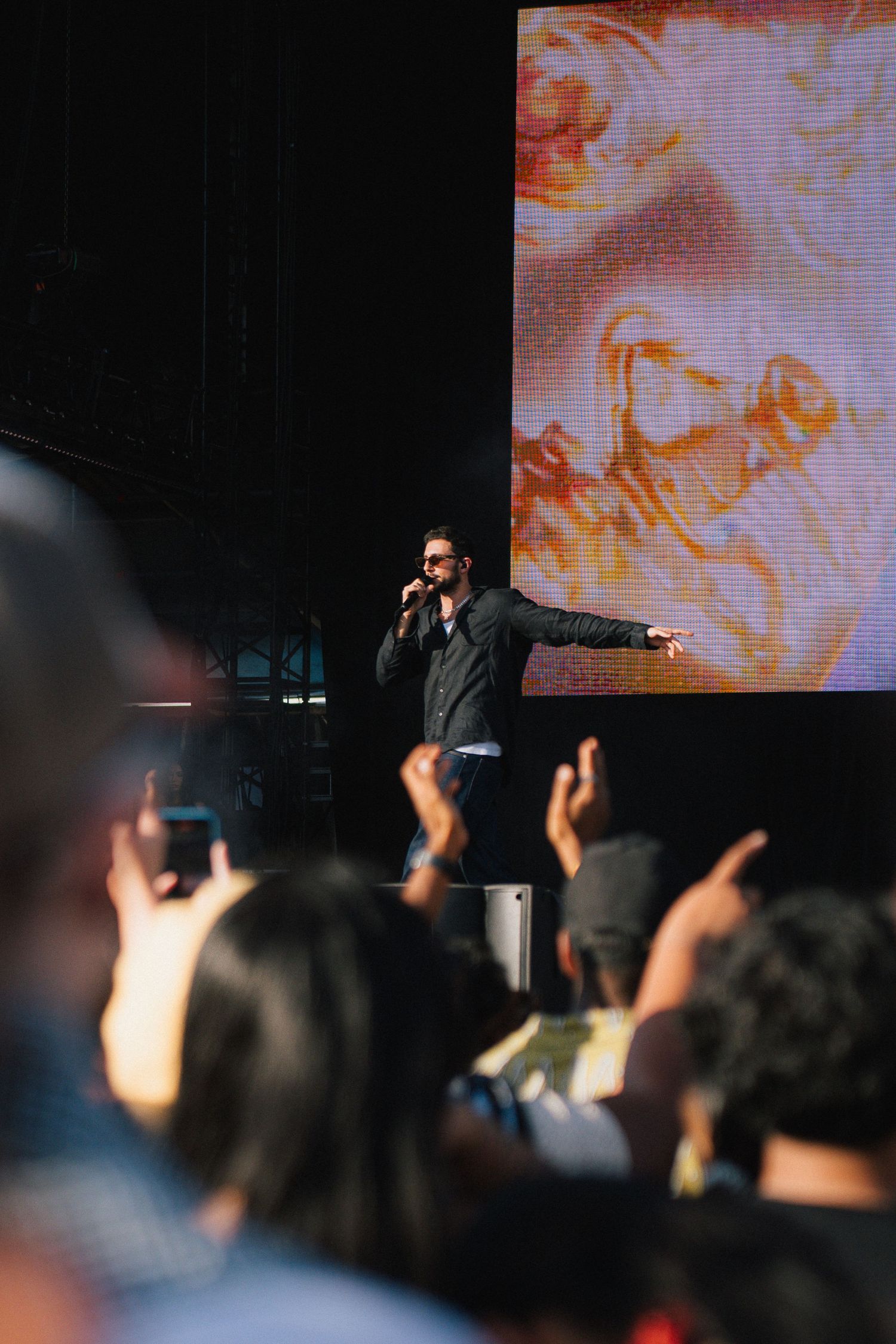 A performer sings on stage with an enthusiastic crowd below and a colorful projected display in the background.