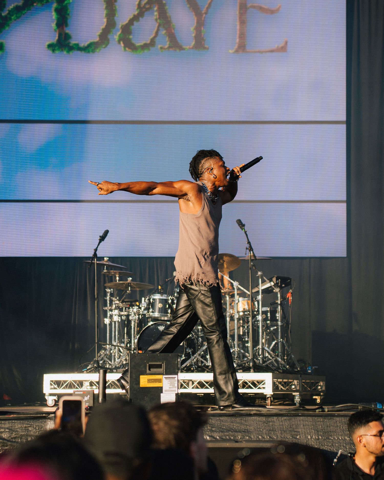 A performer stands energetically on stage with arms spread wide in front of a drum kit and screen displaying text in blue lighting.