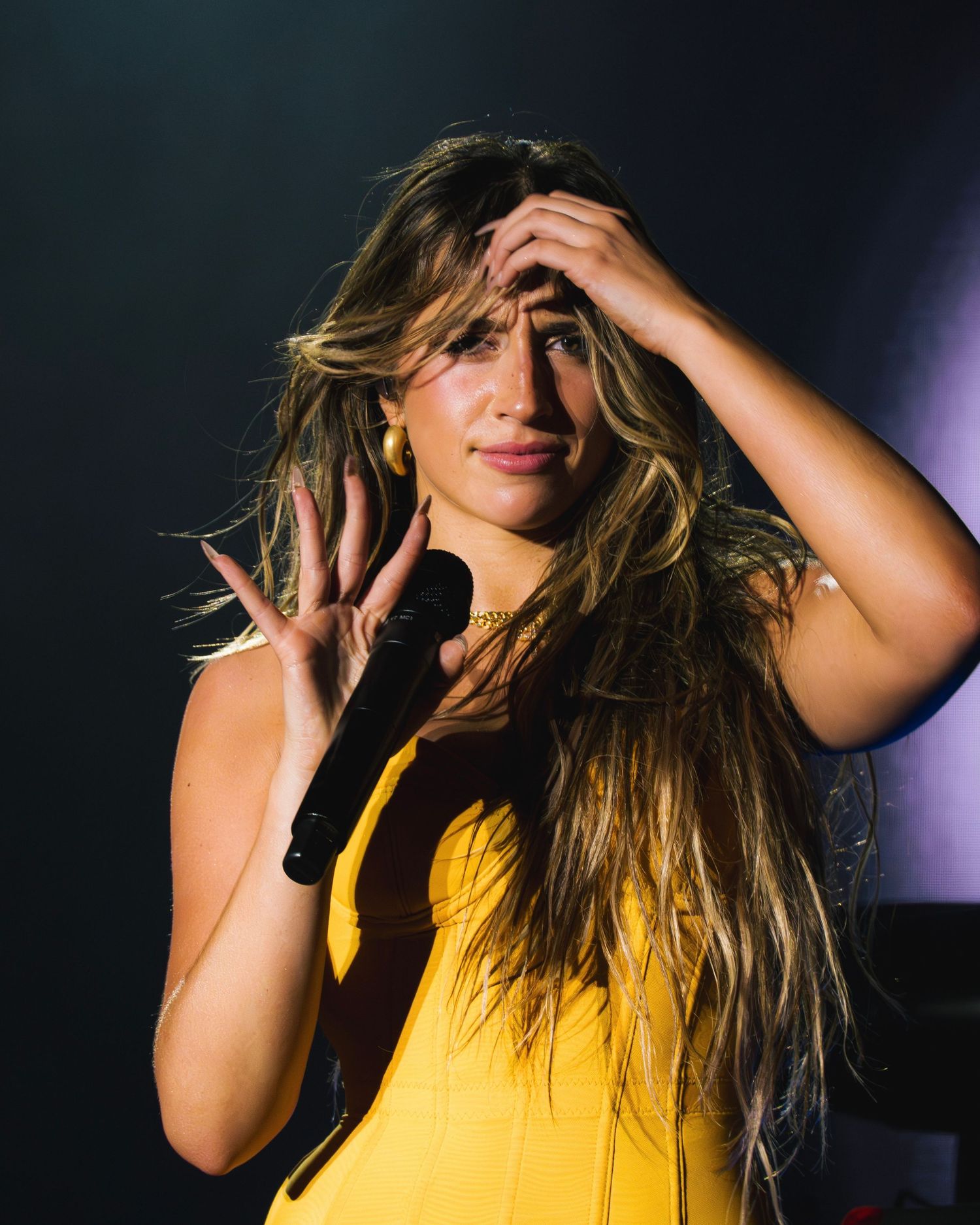 Performer in yellow outfit strikes an animated pose while running fingers through long flowing hair on stage.
