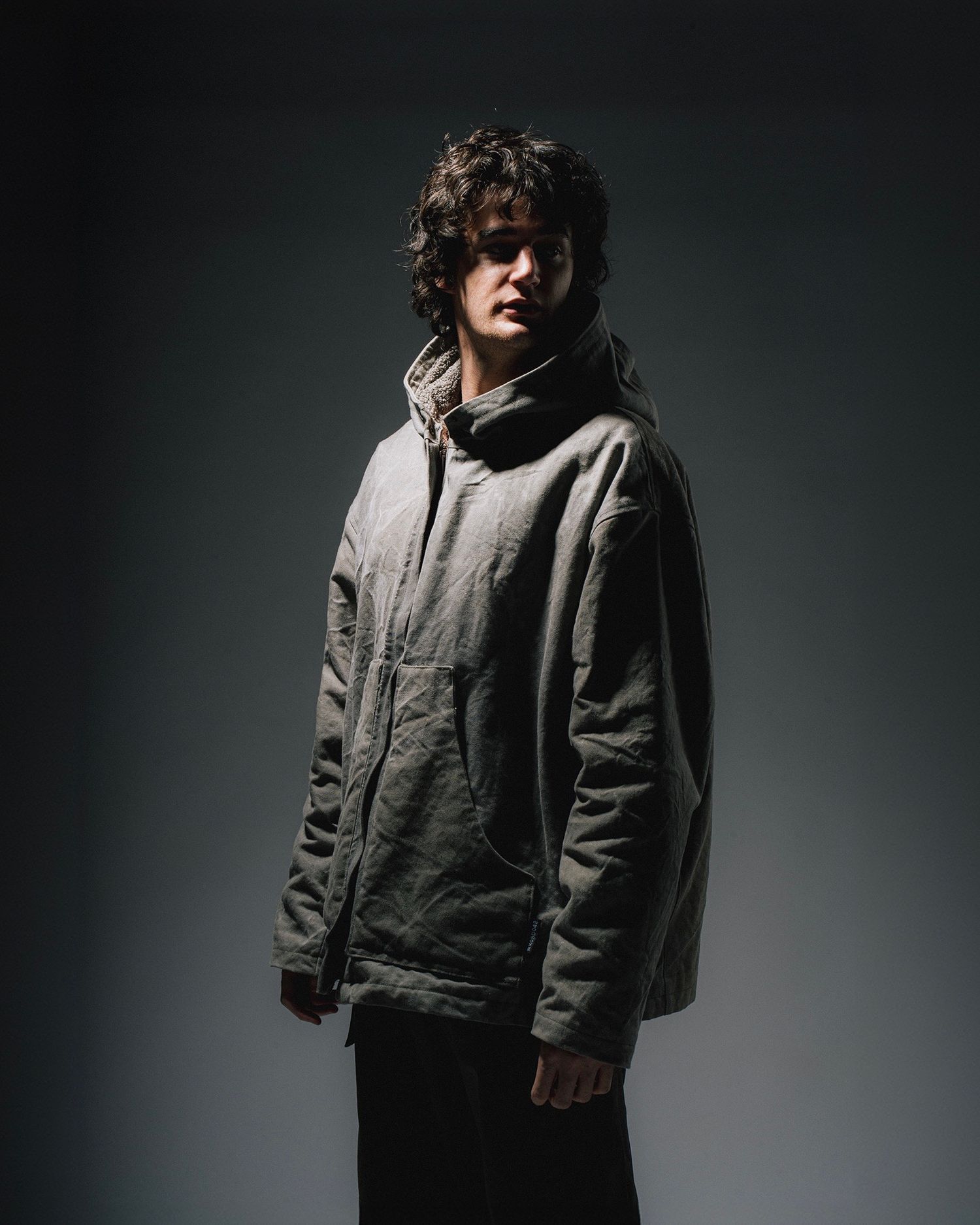 Moody portrait of person in gray jacket against dark background with dramatic lighting.