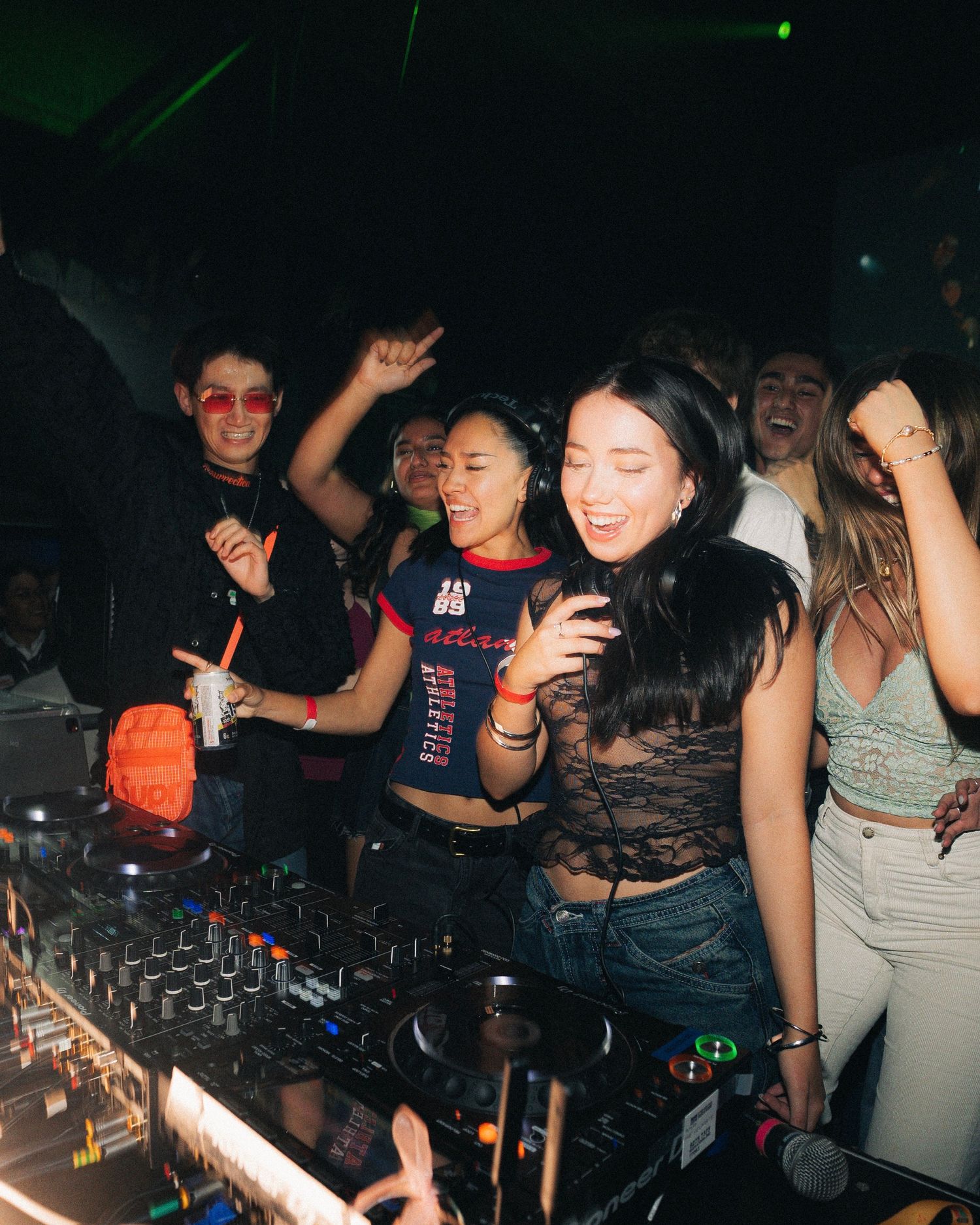 Group of energetic people dancing and performing behind DJ equipment at nightclub.