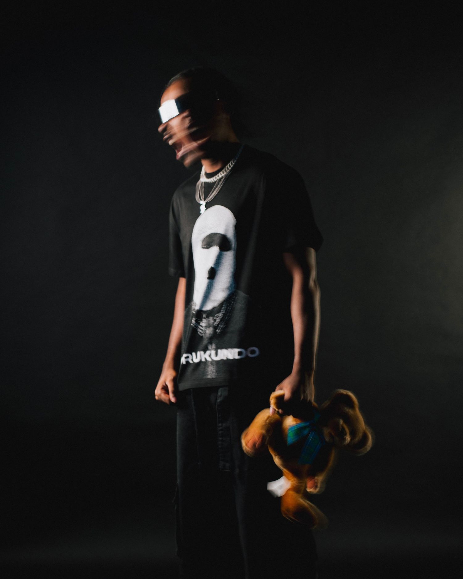 Artistic portrait of person wearing graphic t-shirt and sunglasses holding plush toy against dark background.