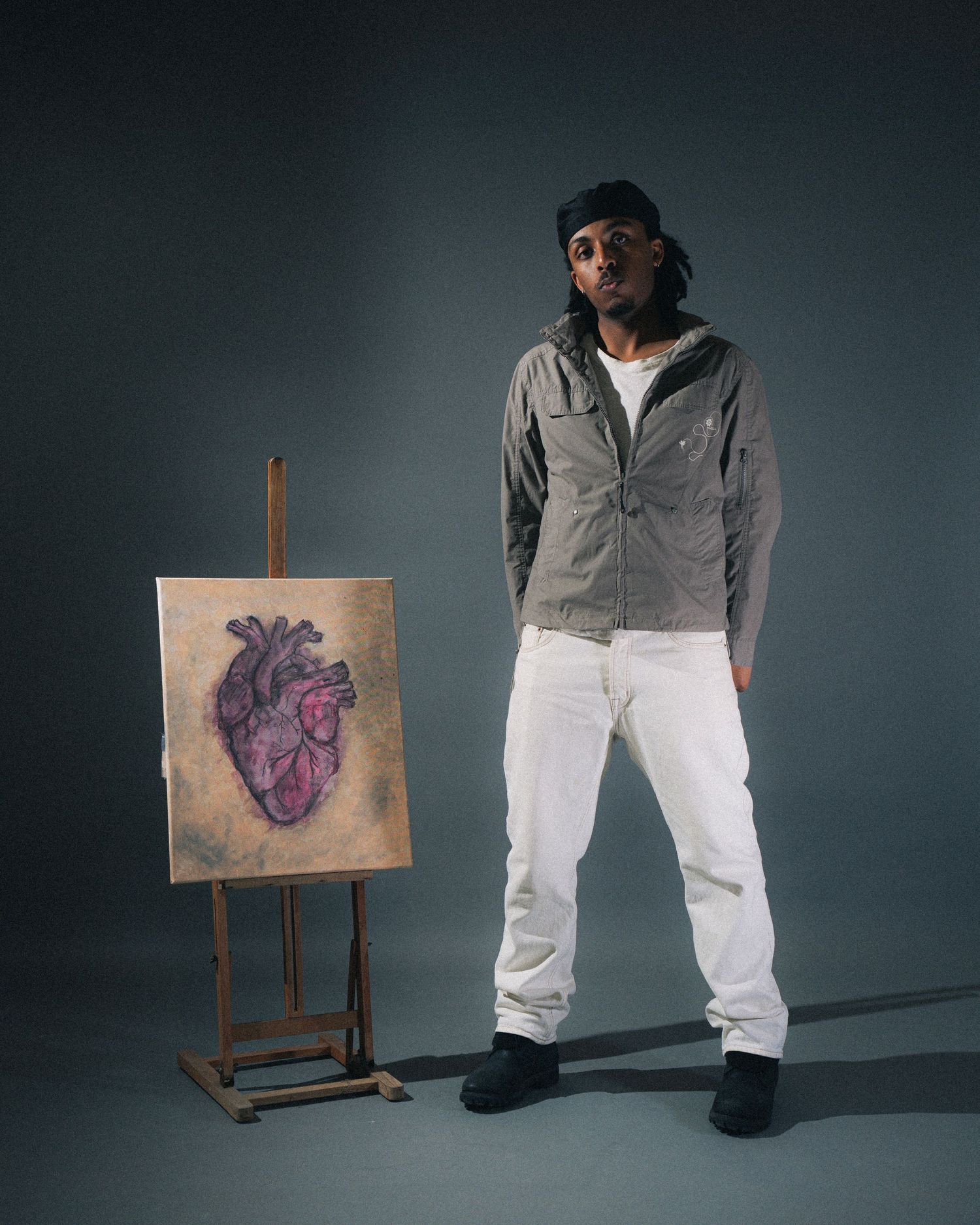 Figure stands next to an anatomical heart painting on an easel against a dark studio backdrop.
