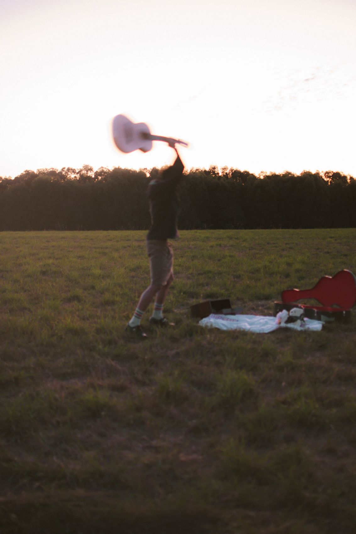 A silhouetted figure holds up a guitar against a sunset sky in an open field with scattered items on the ground.